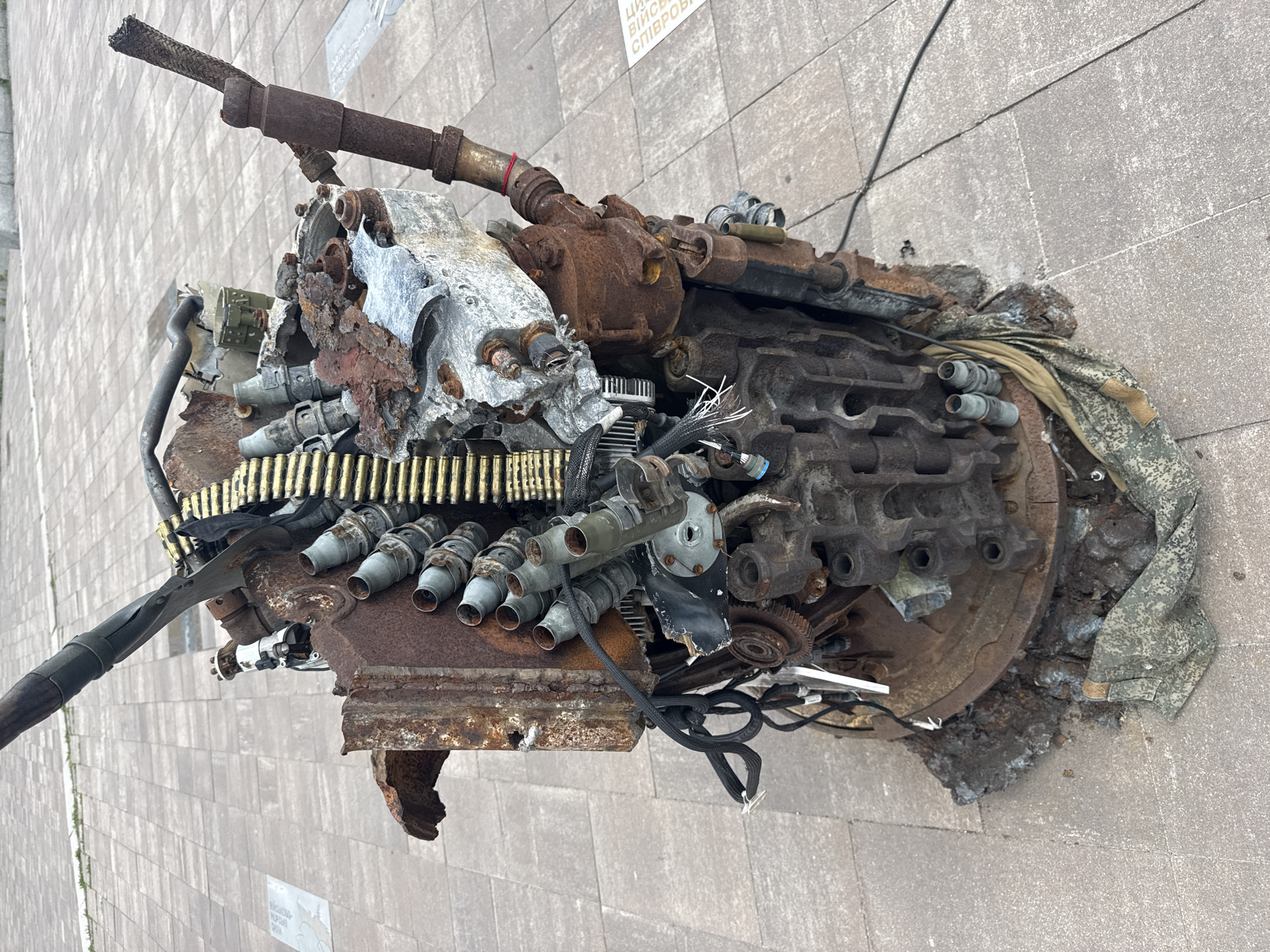 The image shows a sculpture made of destroyed Russian military equipment, displayed on a paved plaza in Kyiv, Ukraine. The sculpture is a complex arrangement of rusted metal parts, including what appears to be a machine gun, ammunition belts, and engine components. Some of the pieces are twisted and damaged, suggesting they were part of a conflict. The sculpture is placed outdoors on a light-colored, tiled surface, with a portion of a building or monument visible in the background. A piece of camouflage fabric is draped around the base of the sculpture. The overall impression is one of a memorial or artistic statement about the impact of war.