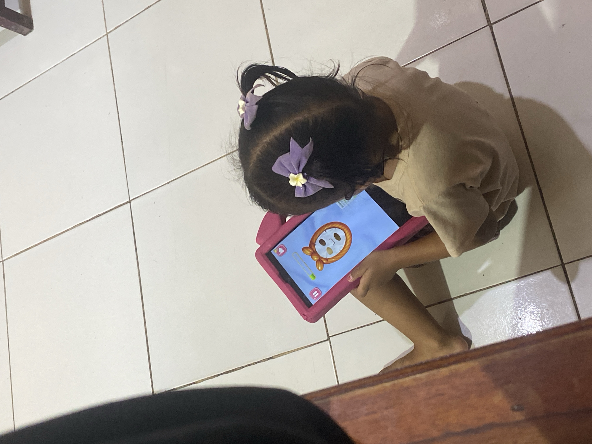 An overhead, high-angle shot shows a young child sitting on a white tiled floor and interacting with a pink tablet. The child, viewed from behind and above, has dark hair styled into two pigtails adorned with purple bows. They are wearing a light-colored, long-sleeved shirt. The tablet displays an image of a cartoon face with a mask. The floor appears to be in a residential setting, possibly a home in Ta', Indonesia, with visible grout lines between the square tiles. A wooden surface, likely a table or furniture, is partially visible in the lower-left corner of the frame, casting a shadow. The lighting suggests an indoor environment, possibly with artificial light sources.