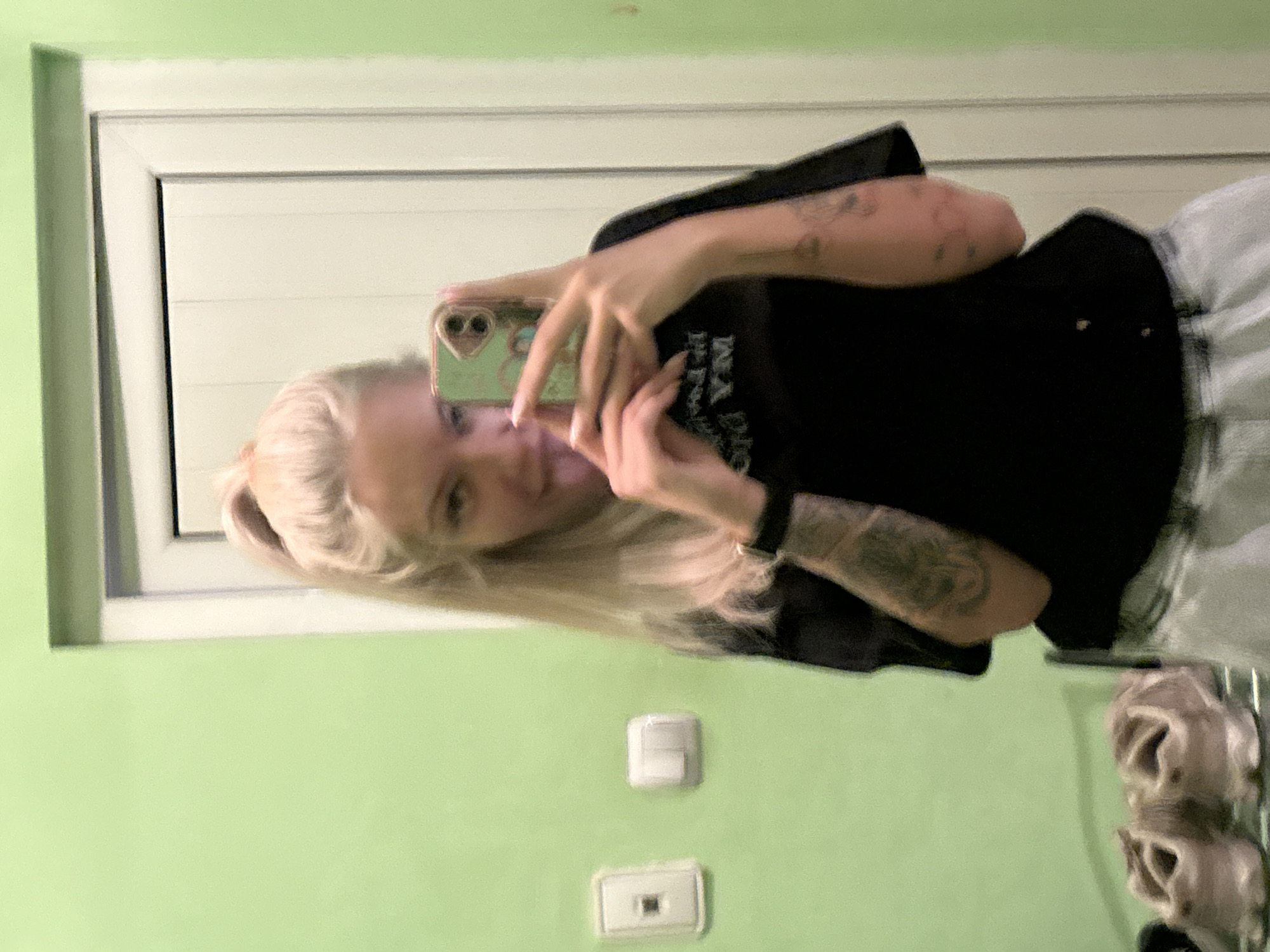 A mirror selfie taken in what appears to be a bathroom or hallway with a pale green wall. The woman in the image has blonde hair pulled back in a ponytail, is wearing a black t-shirt and light-colored patterned pants. She is holding a cell phone up to take the selfie. A white window or door is visible in the background. Below the woman are two light switches, and further down there is a wire rack holding shoes. There is no specific location in Plovdiv, Bulgaria visible in the image.