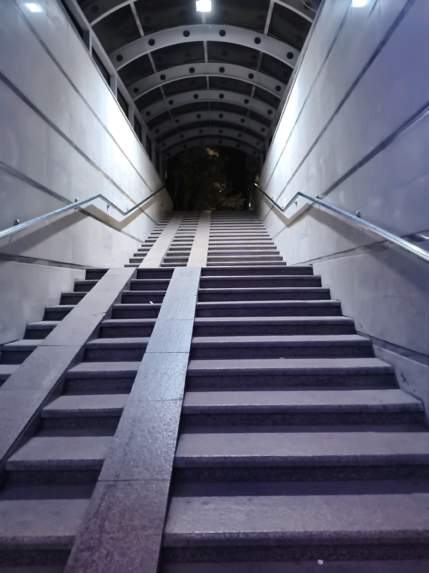 This is a low-angle, full shot of an illuminated pedestrian underpass or walkway in Bishkek, Kyrgyzstan, at night. The structure features a long, arched, metallic ceiling with circular indentations and overhead lighting. Two sets of stairs ascend into the darkness at the end of the underpass, with inclined ramps running parallel to the stairs. The walls are smooth and light-colored, with horizontal lines of trim. Metal handrails run along both sides of the stairs. The foreground shows several wide steps, with ramps positioned on the left and right sides. The lighting creates strong shadows and highlights, giving the scene a somewhat dramatic and modern aesthetic. There are no people visible in the image. The environment appears clean and well-maintained.