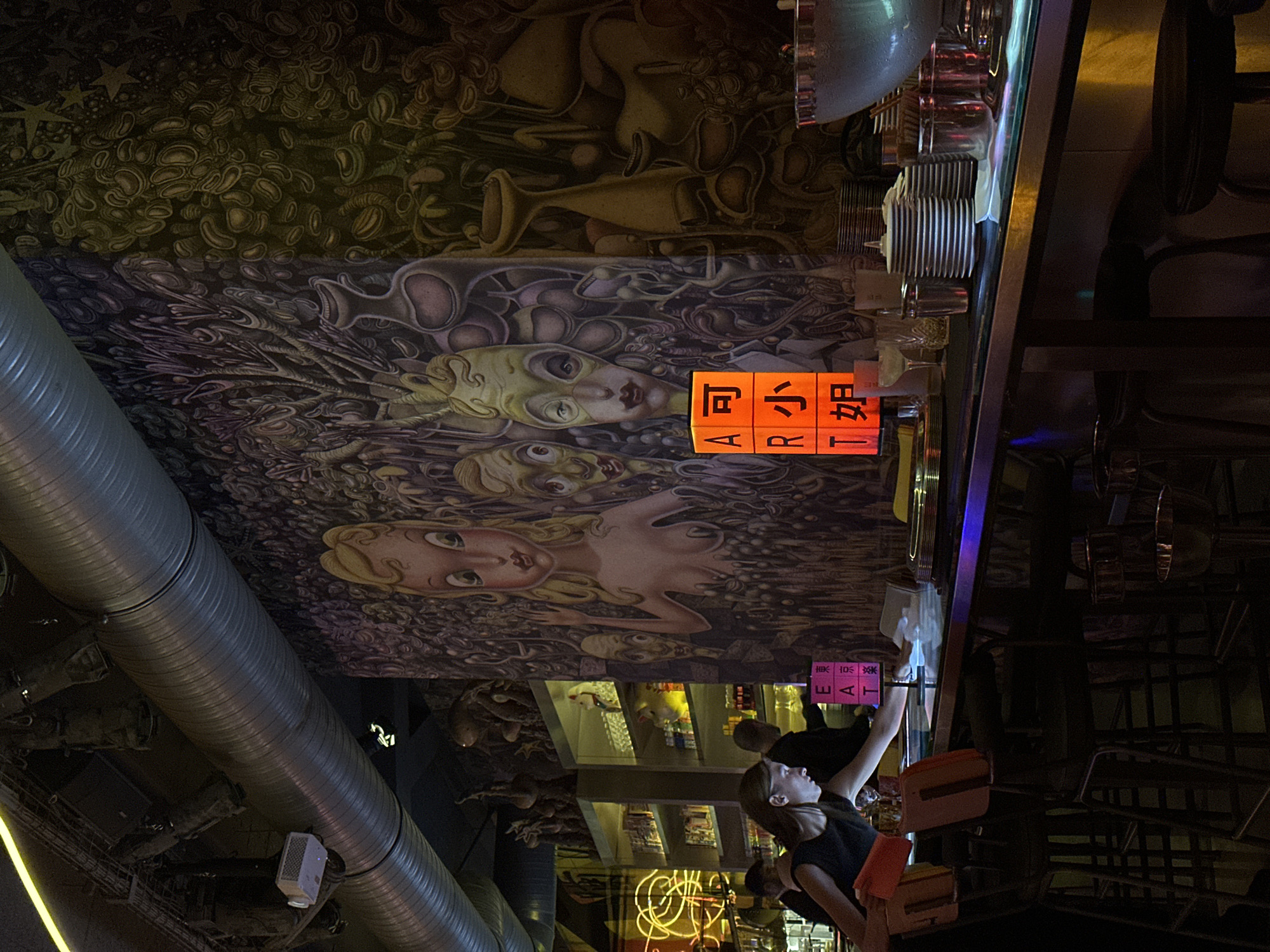 The image depicts the interior of a dimly lit bar with a striking mural covering the back wall. The mural features stylized figures with exaggerated features and a dark, surreal aesthetic. In the foreground, a bar counter is visible, with stacks of plates, glasses, and bar tools. A woman in a black dress is leaning over the counter, seemingly attending to something. Beside her, a cube-shaped light display shows the English letters "E A R T" and Japanese characters. The bar is furnished with tall stools, and overhead, large industrial-looking ducts and lighting fixtures are present. The overall atmosphere is eclectic and moody, suggesting a contemporary establishment. The scene is not definitively located in Paris, France, as no visual cues support such a location.