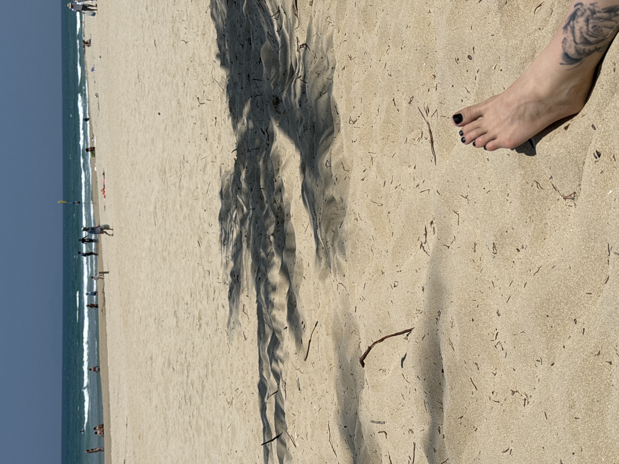 The image depicts a beach scene with a view of the ocean on the left side. The sand is light beige with scattered debris of organic matter such as small twigs and dried grass. Several people are visible in the distance along the shoreline. In the foreground, a shadow falls across the sand, with a person's foot and ankle partially visible on the right. The person has dark nail polish on their toes and a tattoo of a rose can be seen on the ankle/lower leg.
Based on the visible characteristics of the beach, including the wide expanse of sand and the presence of numerous people along the coastline, this scene is likely located on one of the beaches in Đà Nẵng, Vietnam.
