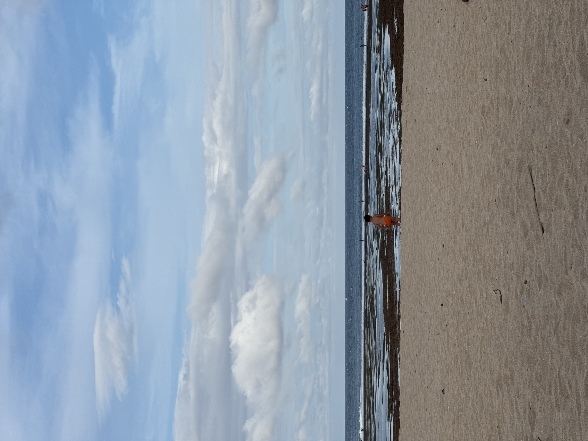 The image captures a beach scene, likely in Kuta Selatan, Indonesia, during daylight. The sky above is a bright blue, dotted with white, puffy clouds. A narrow strip of ocean separates the sky from the sandy beach. The water appears dark, with visible seaweed or algae near the shore. A person, possibly a child, wearing orange clothing is standing at the water's edge. The beach itself is a light tan color, with various small objects scattered across the surface. The image is vertically oriented.