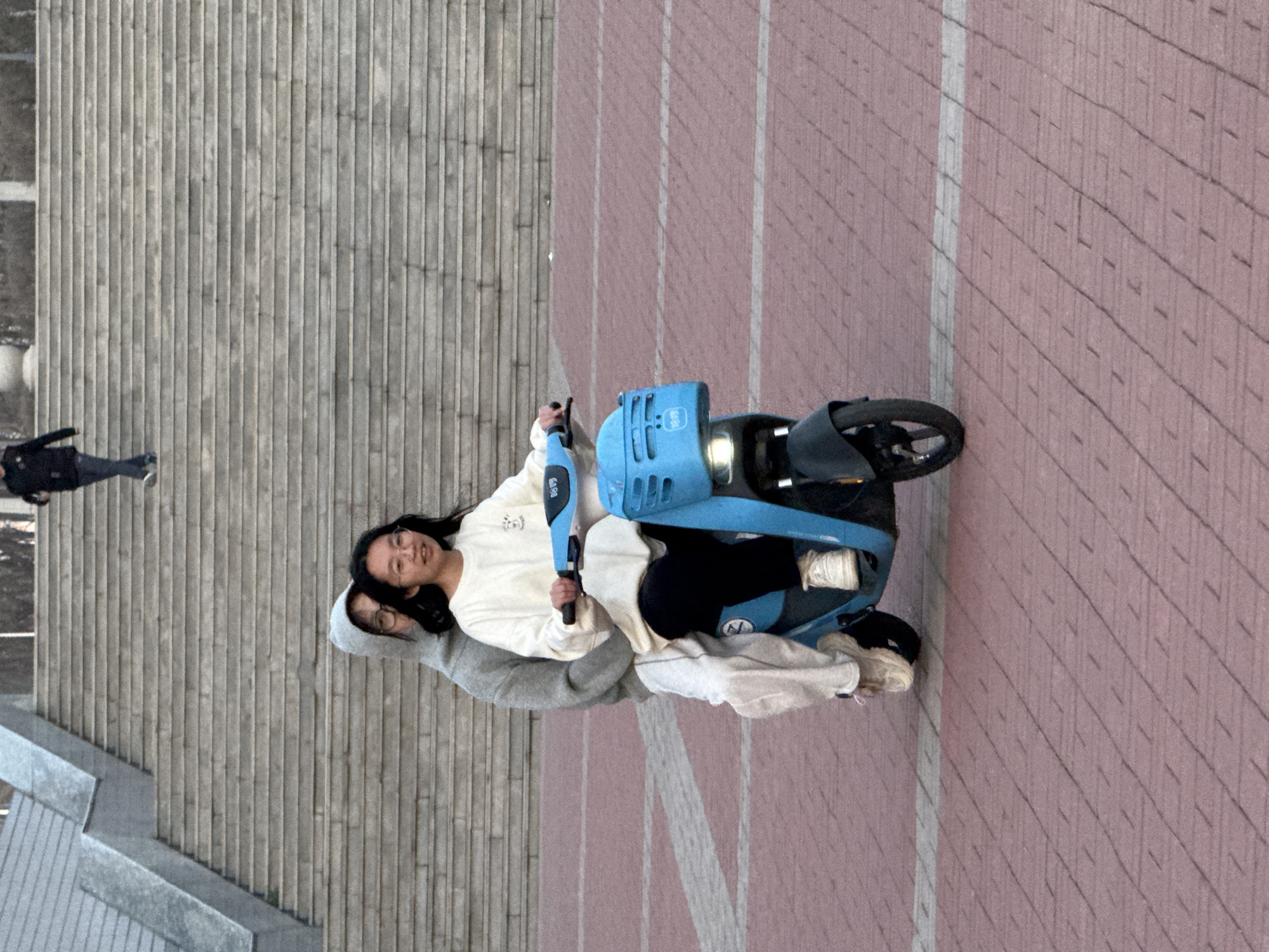The image shows two individuals riding a blue electric scooter on a brick-paved walkway. One individual is driving the scooter, and a second individual is riding as a passenger behind the driver. Both people appear young. The scooter has a light in the front. The background includes a set of stairs with vertical concrete paneling and a person walking near the stairs. The scene's location is Zhongtun, China.