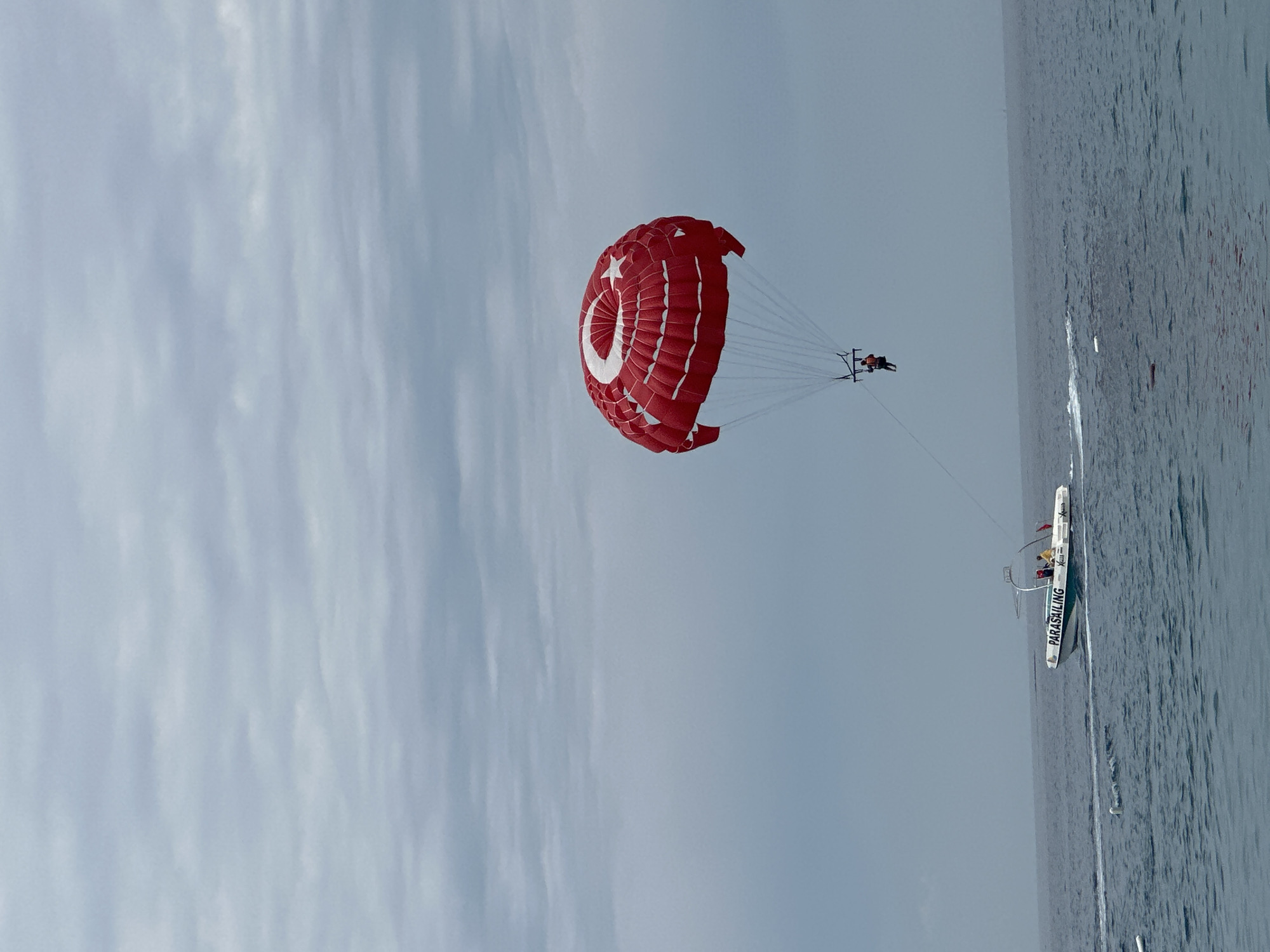 The image captures a vibrant parasailing scene over a body of water, likely the sea, under a cloudy daytime sky. A large, bright red parasail dominates the upper center of the frame, featuring a prominent white crescent moon and star symbol, indicative of the Turkish flag. Two individuals are visible, suspended in a harness beneath the parasail, enjoying the aerial view.
The parasail is being towed by a white motorboat, visible on the water to the right. The boat has the word "PARASAILING" clearly written in white on its side, confirming the activity. Several people are also discernible on board the boat. A thin rope connects the parasail to the boat, extending diagonally across the frame.
The water below appears calm with subtle ripples, reflecting the muted light from the overcast sky. The sky itself is largely covered by a uniform layer of light grey clouds, suggesting either an overcast day or a period of high cloud cover. The overall impression is one of leisure and outdoor recreation in a coastal setting, consistent with the location context of Fethiye, Turkey.
