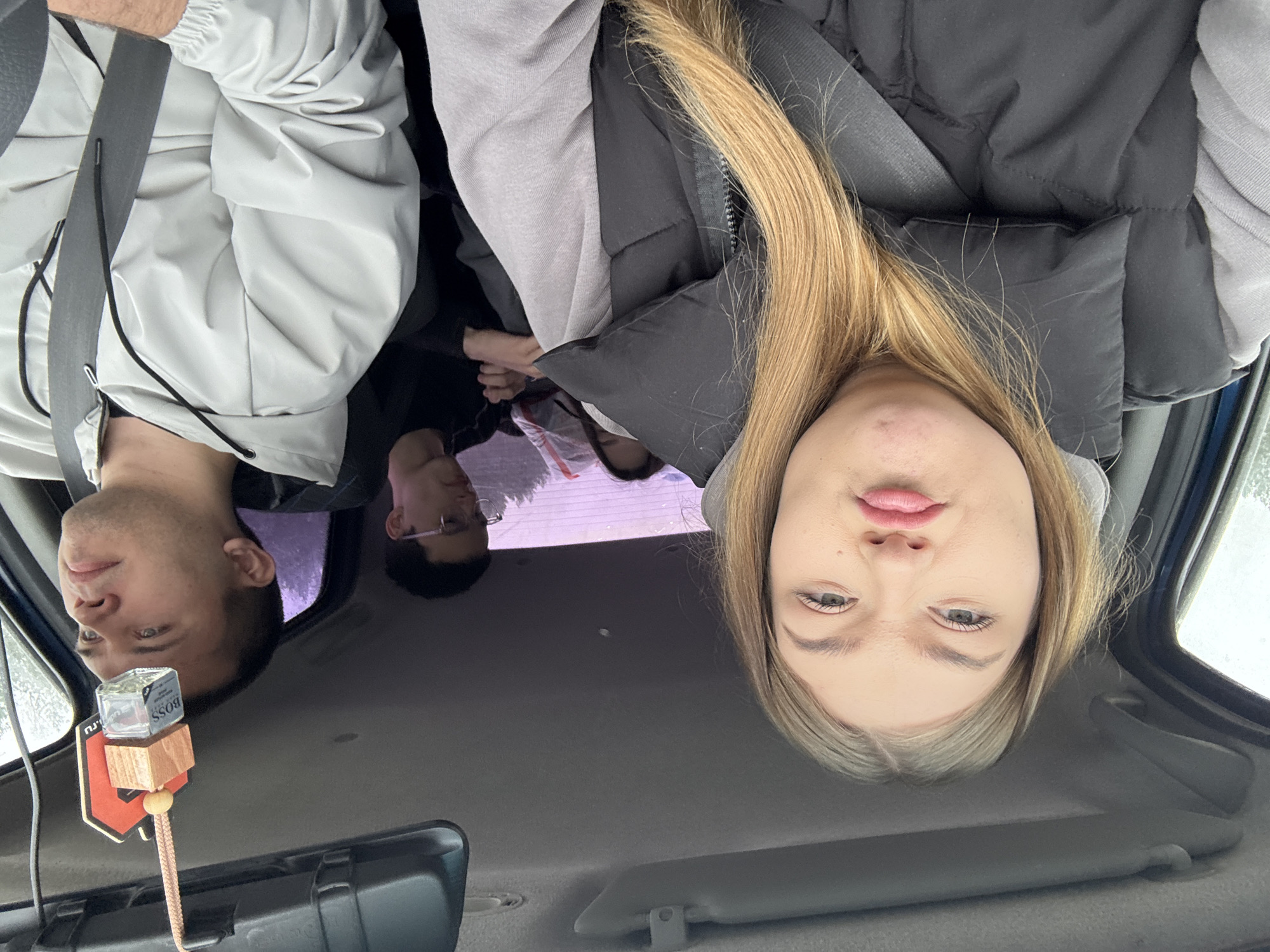The image shows the interior of a vehicle, with four individuals visible. In the front passenger seat, a blonde woman is wearing a black vest and grey hooded sweatshirt. She is positioned close to the camera, partially obscuring the view. In the driver's seat, a man is wearing a light grey jacket and a seatbelt. Behind them, in the back seat, two other individuals are present. One is wearing glasses and a black shirt. A car air freshener is hanging from the rearview mirror, with the "BOSS" logo visible on the bottle. The location is not discernible as being within the city of Maksimovo, Russia.