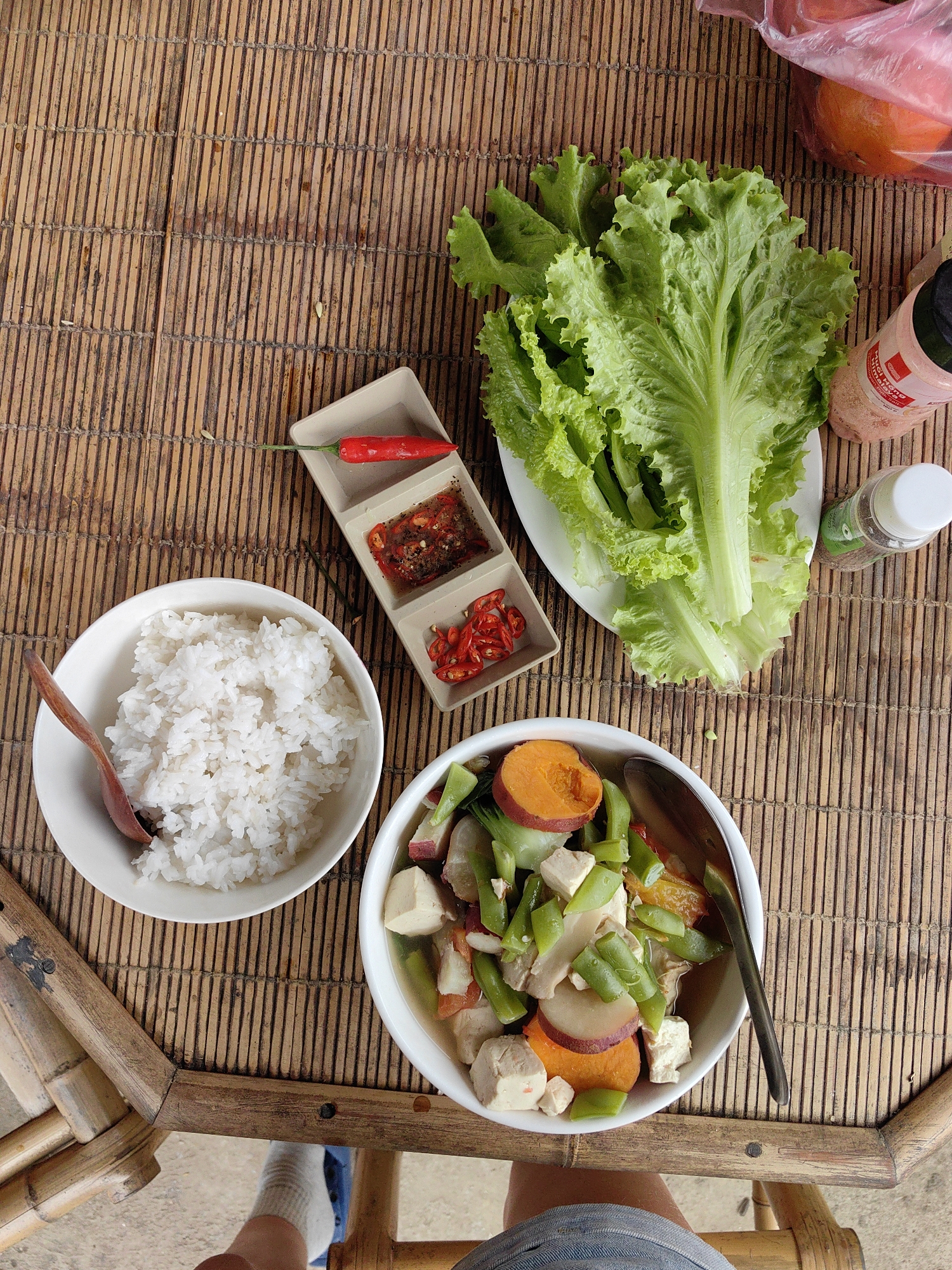 The image shows a meal set on a woven bamboo table, likely in a rural or outdoor setting, potentially in or near Xã Đàm Thủy, Vietnam, given the regional style of the food and woven table.
Food items include:
- A bowl of white rice with a wooden spoon.
- A bowl of vegetable stew containing green beans, tofu, and orange slices, with a metal spoon.
- A plate of leafy lettuce.
- A small, sectioned dish holding a red chili pepper, chili paste, and chopped chili peppers.
- Two bottles of condiments.
Other details:
- A bamboo chair frame is partially visible.
- A bag of fruit, possibly oranges or mandarins, is visible in the background.
- Part of a person's leg, wearing denim shorts and sandals, is visible.