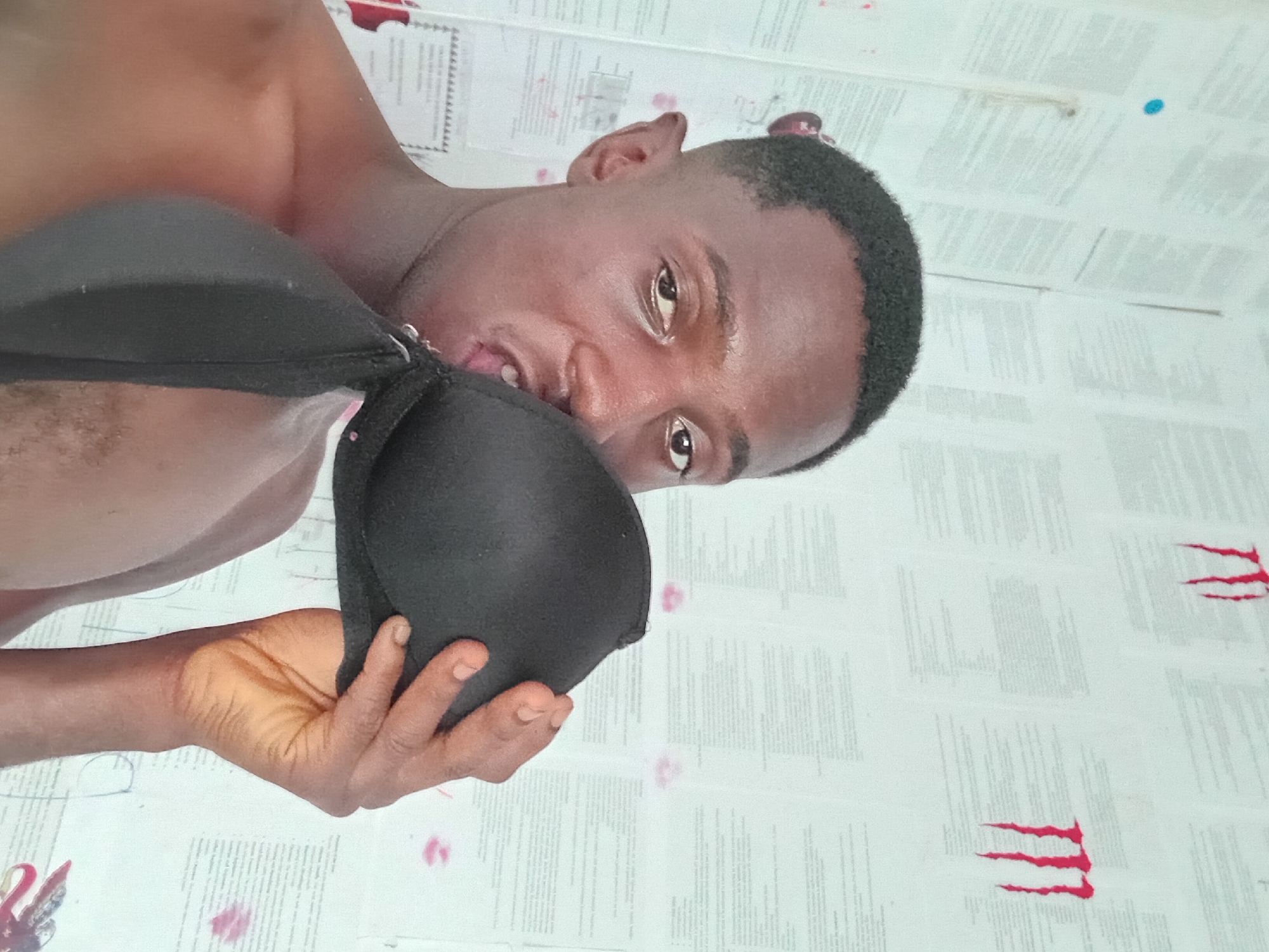 A topless man is holding a black bra with his left hand and putting it in his mouth. The background is a white surface covered in text. There are red claw marks at the corners of the image. The location is not evident from the image.