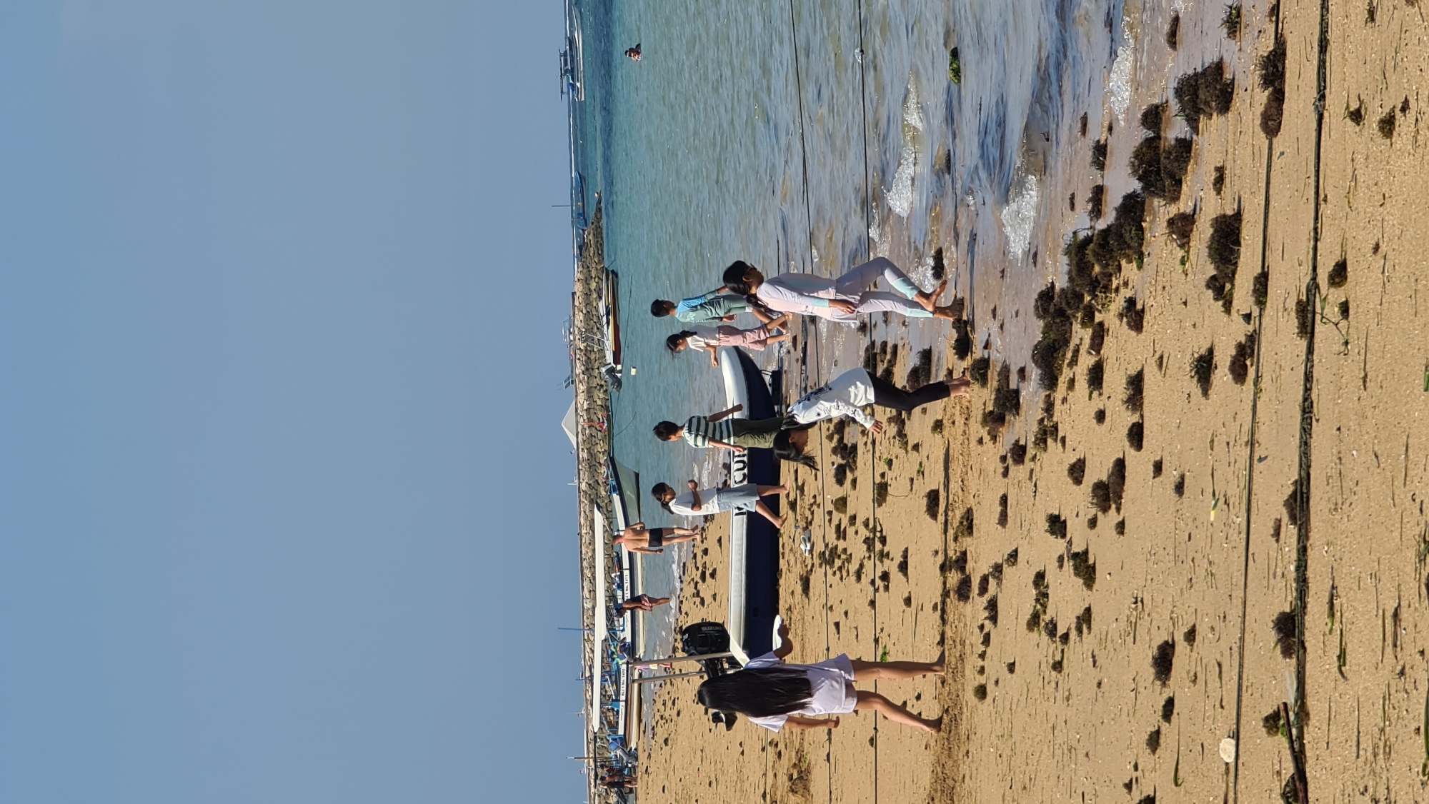 The image depicts a beach scene in Denpasar, Indonesia, likely on a sunny day with clear skies. The foreground is dominated by a sandy beach strewn with small clumps of dark seaweed. Several children are visible, scattered across the beach and at the edge of the water.
In the mid-ground, a long, blue and white inflatable boat is partially beached on the sand, with the word "GU" partially visible on its side. Several other boats, including what appear to be traditional Indonesian jukung boats with outriggers, are moored in the shallow turquoise water behind the inflatable boat. A stone breakwater stretches across the background, with more boats anchored beyond it.
On the beach, a young girl with long black hair is standing in a light purple dress, facing left and holding a white plate. To her right, another young girl in a white top and black pants is bending down, appearing to examine something on the sand. Further back, a group of children are walking towards the water or playing near the boats. One boy in a green and white striped shirt is looking down. A girl in a pink and white patterned outfit is walking near him, and another girl in a light blue long-sleeved top is behind her. A man in swim trunks walks away from the water in the background. In the far distance, a person is visible swimming in the ocean. The overall atmosphere is one of a relaxed day at the beach.