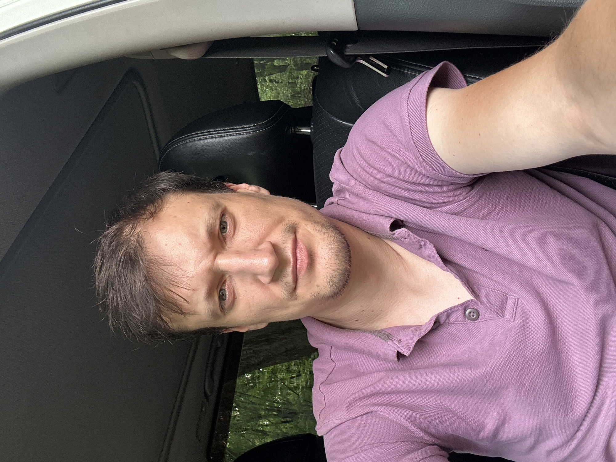 A man with brown hair and a light complexion is seated in the driver's seat of a vehicle. He is wearing a purple polo shirt. The interior of the car, including black leather seats, is visible. Through the windows, a wooded area with green trees can be seen, suggesting an outdoor, possibly rural or suburban, setting outside of Shatava, Ukraine. The man is looking at the camera, and his expression is neutral.