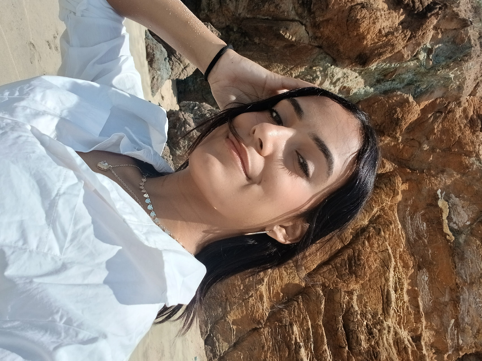 This close-up image features a young woman lying outdoors, posing for a photograph in Pedro González, Venezuela. She looks up towards the camera with a soft, relaxed smile. Her dark hair is parted, with some strands falling across her face. She has dark eyes and a clear complexion, with distinct shadows from bright sunlight accentuating her features.
The woman wears a white collared shirt with long sleeves. Around her neck is a silver necklace featuring small, heart-shaped charms and a larger central pendant. A thin, gold-colored earring is visible in her right ear, and a black hair tie adorns her left wrist, which is raised and positioned near her head.
The background on the left shows sandy ground, while the right side and upper portion of the frame are dominated by a large, textured rock formation. These rocks display various earthy tones of brown and orange, with some areas showing lighter greenish-grey patches, indicative of natural weathering or mineral deposits. The bright, direct sunlight suggests the photo was taken during the day under clear skies. Her expression conveys a sense of ease and contentment. No text is visible in the image.