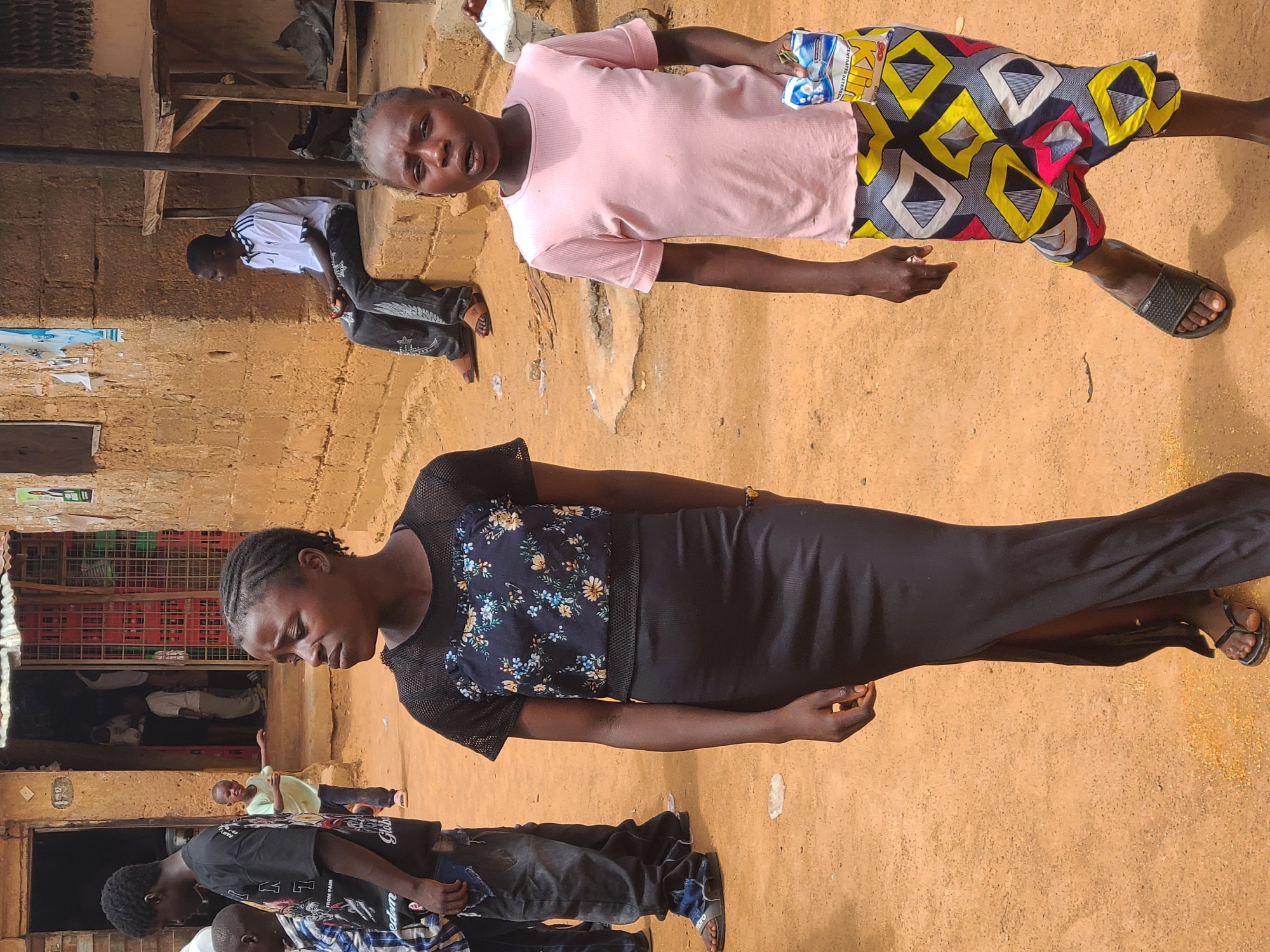 The image shows two women standing outside a building in Unknown, Unknown. One woman is wearing a pink shirt and patterned shorts, while the other is wearing a long black dress with a floral top. Other people are visible in the background. The ground is dirt.