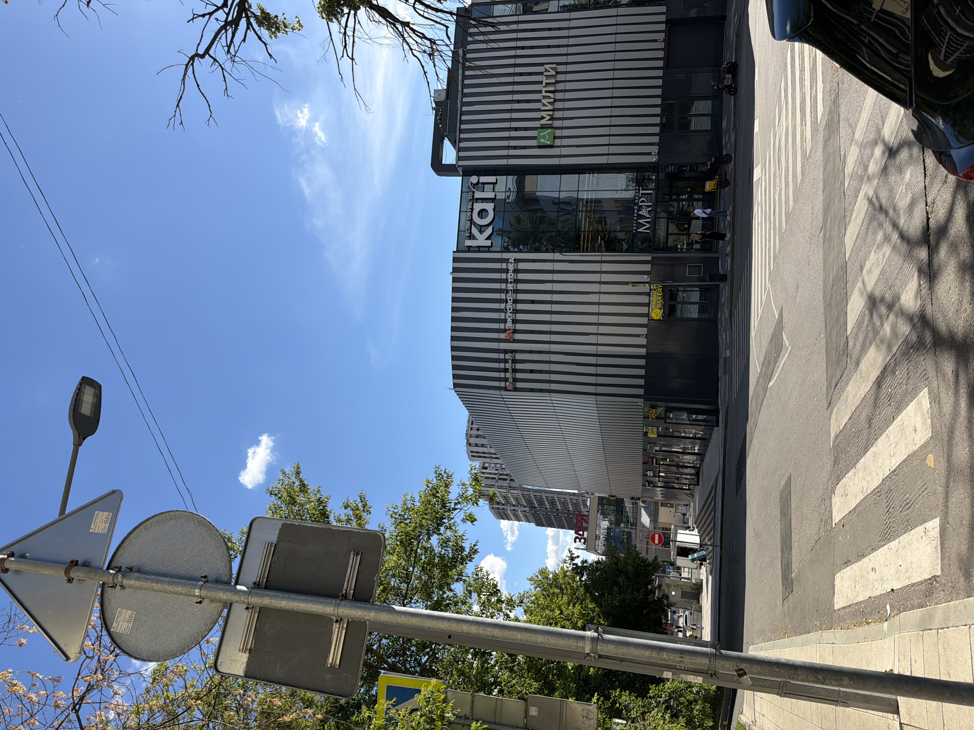 The image depicts a street scene in Moscow, Russia, on a bright, sunny day. The sky is a clear blue with a few scattered white clouds. The camera is angled upwards, with a tree branch and power lines visible in the foreground against the sky.
Dominating the scene is a modern, multi-story building with a distinctive facade of horizontal black and white stripes. Several large signs are visible on the building. One prominent sign reads "Kari" in large, stylized letters, suggesting a shoe store. Another sign displays "МАРТ" (MART) and "ТОРГОВЫЙ ЦЕНТР" (Shopping Center), indicating a commercial establishment. Above these, a sign with a green logo and the text "Мильти" is also visible. Further down, a yellow sign with the text "BAKERY" suggests a bakery on the ground floor.
In front of the building, a pedestrian crossing with white stripes is clearly marked on the asphalt road. A dark-colored car is partially visible on the right side of the road, with its reflection on the wet asphalt. A street light and several traffic signs, including a triangular warning sign and circular signs, are mounted on a metal pole in the lower left portion of the image.
People are visible in the scene, though small in scale. One person stands near the entrance of the "MART" shopping center, and others appear to be walking in the vicinity. The overall impression is of a bustling urban environment, likely during daytime given the bright sunlight and clear skies. The architectural style of the building and the Cyrillic text on the signs firmly place the location in Russia.