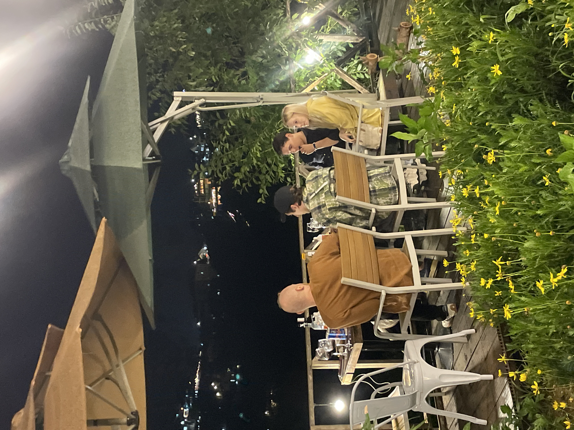 This image captures a nighttime scene at a restaurant or cafe in Da Lat, Vietnam, with a panoramic view of the city lights in the background. The foreground features a wooden deck adorned with lush green vegetation and small yellow flowers. Four individuals are seated at a wooden table under a large, two-tiered green umbrella.
A bald man in an orange sweatshirt is seated at the table, facing away from the camera, with his back to the viewer. To his right, a person wearing a plaid shirt and a black baseball cap is also seated at the table, facing partially towards the right. Further to the right, a man in a dark shirt is seated, leaning forward and appearing to converse with a woman beside him. The woman has blonde hair and is wearing a yellow hooded jacket.
On the table are various items including glasses, bottles, and what appears to be a can. Two white metal chairs are positioned at the table, one to the left of the bald man and another to the right of the woman in yellow. A second table is partially visible to the left, with a white metal chair in front of it. The overall ambiance suggests a relaxed evening gathering.