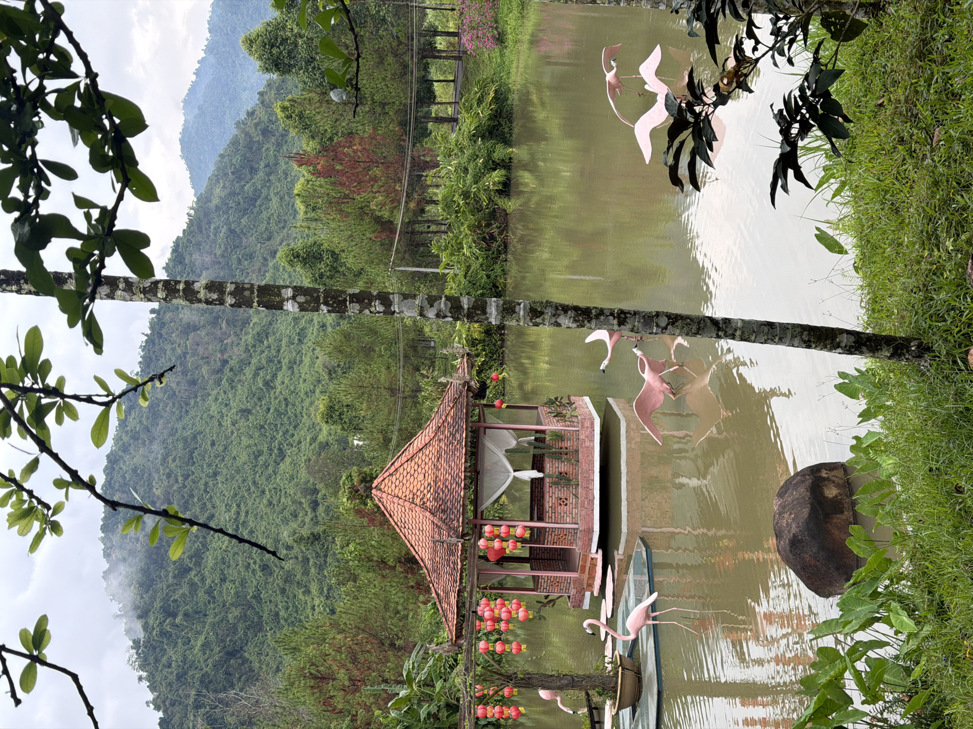 The image depicts a serene landscape in Khanh Vinh, Vietnam. A misty mountain range is visible in the background. In the foreground, a body of water reflects the sky and surrounding foliage. A small, ornate pavilion with a red-tiled roof sits at the water's edge. Pink flamingo statues are scattered around the pavilion and in the water. Lush vegetation, including trees and grass, frames the scene. The image has been rotated ninety degrees counterclockwise.