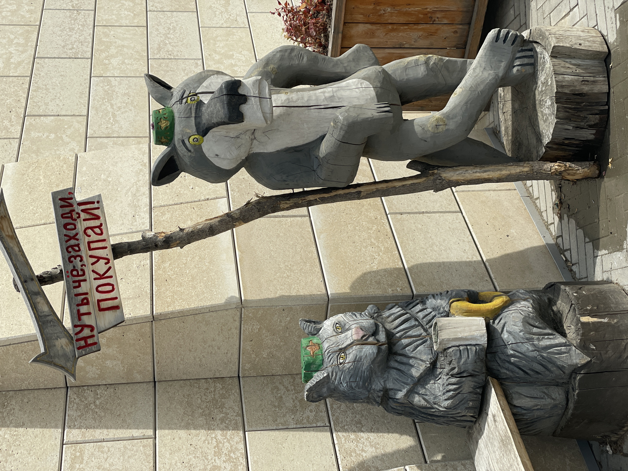 The image captures two wooden sculptures of wolves in Kazan, Russia, placed outside a building with a light-colored tiled facade. The lower wolf, appearing more like a bear, is on the ground, propped against a section of tree trunk, with a small wooden sign in its mouth. The upper wolf reclines horizontally on another section of tree trunk. An arrow-shaped wooden sign, with Cyrillic text in red, protrudes from the wolf's back, indicating the direction for customers. The scene suggests a playful and welcoming entrance, likely for a shop or establishment, using local folk art imagery. The location is assumed to be in Kazan, Russia due to Cyrillic writing and location, which is often associated with that city.