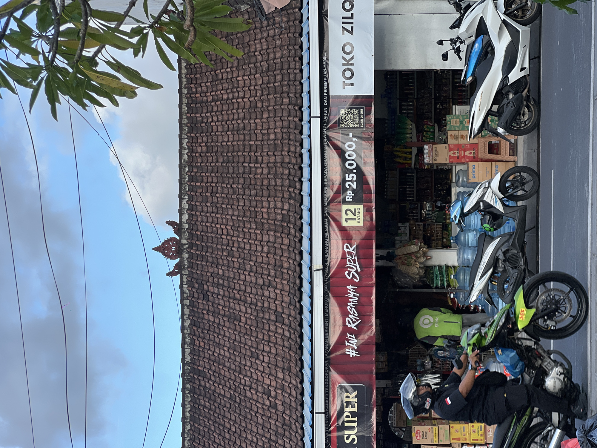 The image depicts a street scene in North Kuta, Indonesia, showcasing a small shop identified as "TOKO ZILO." The shop's facade is partly obscured by promotional banners. Parked along the street in front of the shop are several motorcycles. One rider is adjusting gear. The background includes a building with a tiled roof. The sky is partly visible above the buildings and trees.