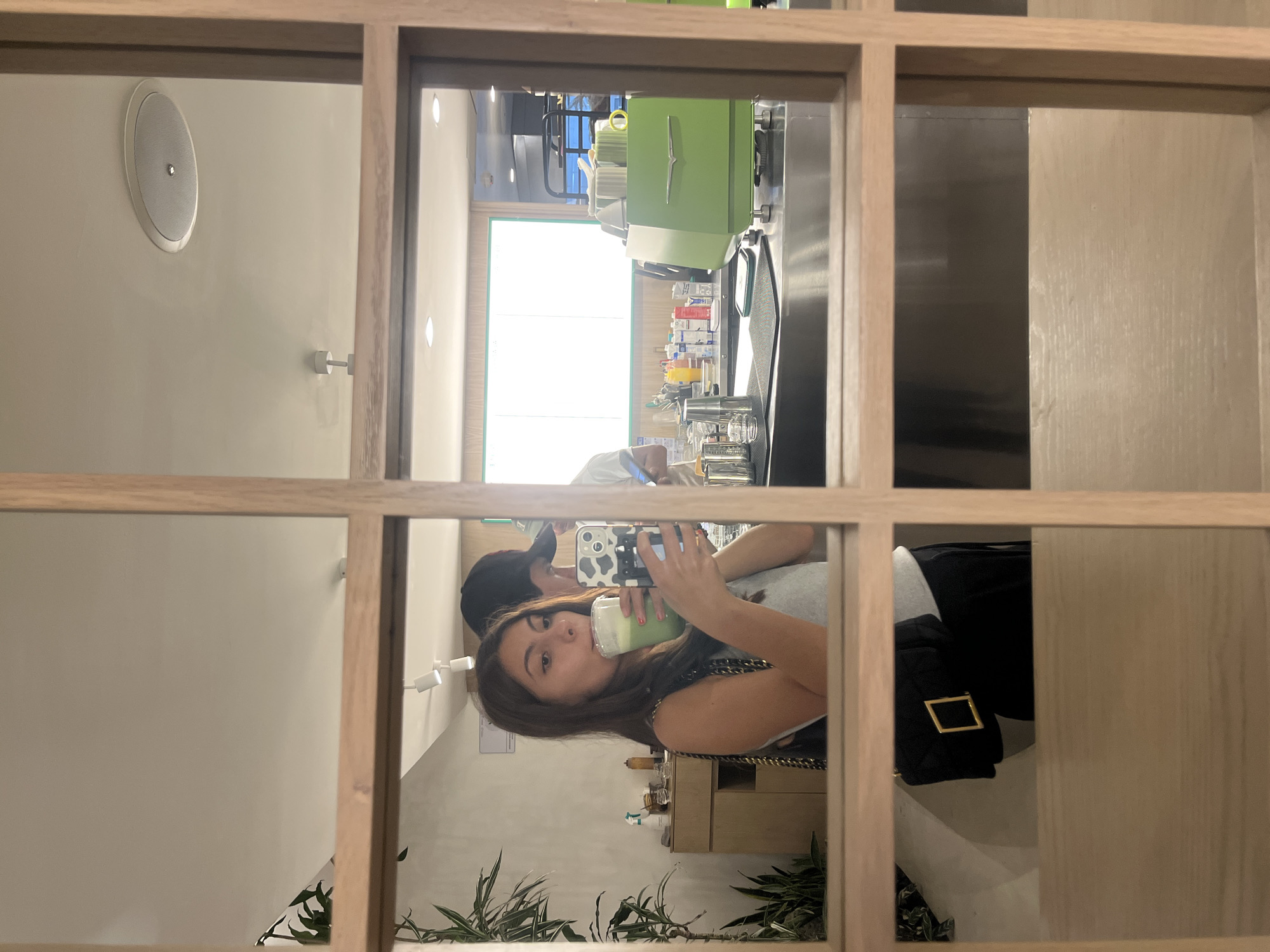 The image shows a reflection in a multi-pane glass frame, capturing a scene inside what appears to be a café or restaurant. A woman is holding a drink, possibly a matcha latte, and taking a selfie with her phone. A man stands behind her, partially visible. The background features a coffee or drink preparation station, including a bright green espresso machine and various bottles of ingredients. The interior design is modern and minimalist. Plants are visible at the bottom of the frame. Based on the overall style and known locations, it is suggested the location is within the city of Puebla, Mexico.