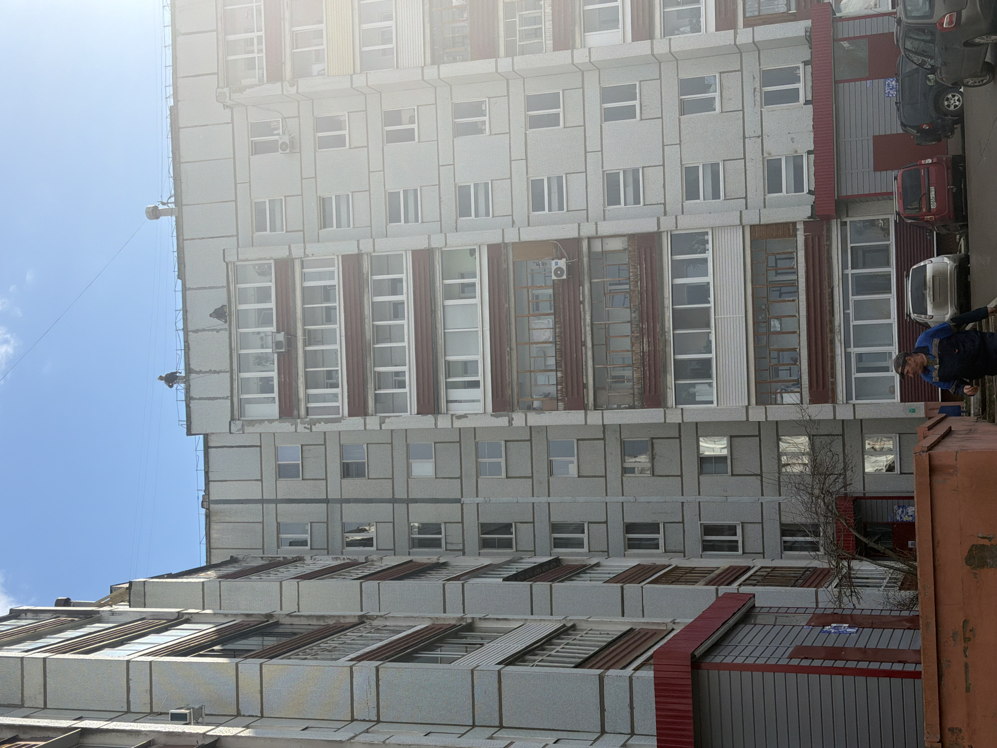 This bright, sunny daytime image captures an urban scene in Chaikovsky, Russia, dominated by a large, multi-story building. The building features a grey and white paneled facade with numerous windows and enclosed balconies, accented by vertical reddish-brown elements. Several air conditioning units are visible on its exterior walls.
On the roof of the tall building, two figures, appearing to be industrial climbers or maintenance workers, are present, possibly engaged in preparations or work, with ropes visible. At ground level on the right, a man in a blue work uniform stands, observing the scene.
Adjacent to the main building is a lower structure with a similar architectural style, featuring a roofline with prominent red-colored accents and horizontal window designs. A street or driveway runs in front of these buildings, where multiple cars are parked. A red car in the foreground prominently displays a Russian license plate, "M004YH 59". A dark SUV is parked further down.
The sky is clear and bright blue, suggesting good weather. A bare tree stands next to the lower building on the right. The architecture is characteristic of functionalist apartment blocks common in Russian urban environments. Some indistinct signs or posters are visible on the walls of the lower buildings.