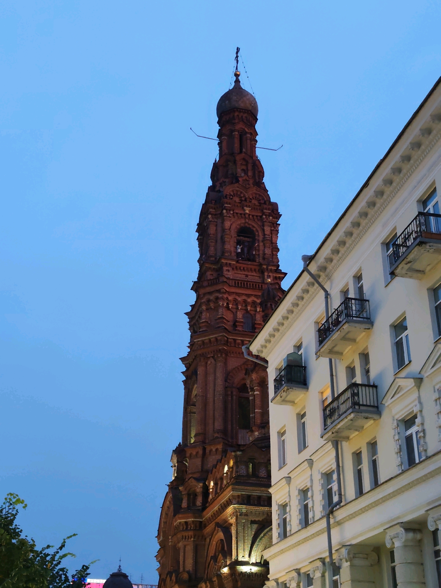 The image shows the Kul Sharif Mosque minaret and a white building in Kazan, Russia. The minaret is tall, made of red brick, and has intricate architectural details. It is topped with a golden dome and a crescent moon. The white building has balconies with black railings and arched windows. The sky is a clear blue, suggesting it is either early morning or late evening. The lighting indicates a twilight setting, with some artificial lights visible on the buildings. There are no people or animals visible in the frame. The scene is a blend of historical architecture and a modern urban environment.