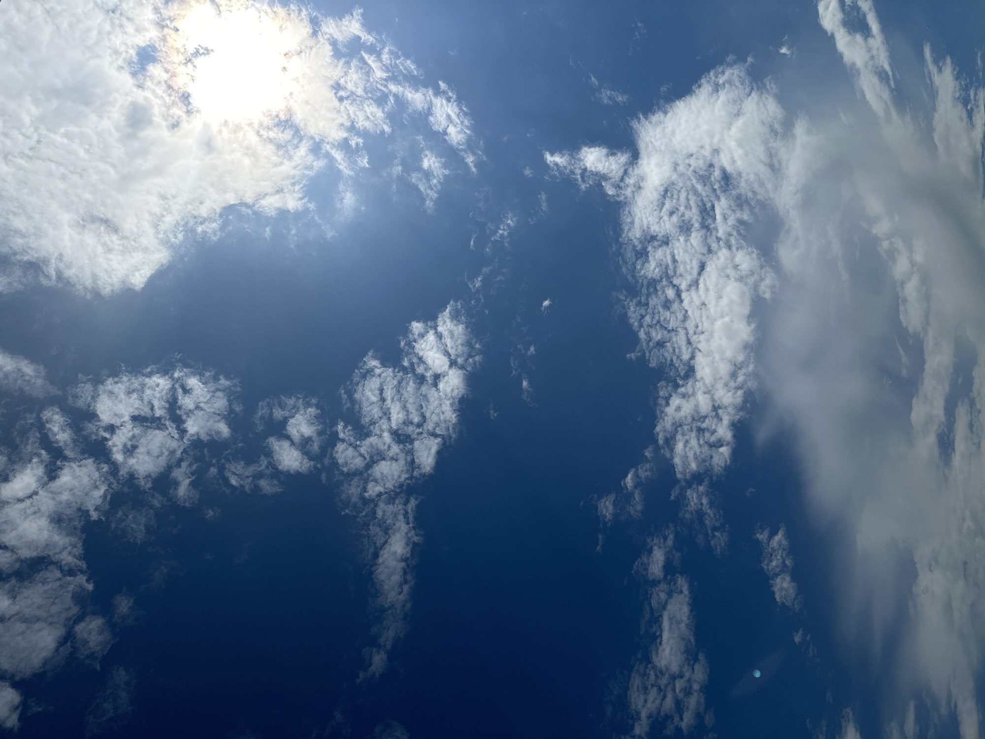 The image shows a bright blue sky with scattered white clouds on a sunny day. The sun is partially obscured by clouds in the upper right portion of the image, casting a bright glare and creating lens flare. There are no people, subjects, or specific objects visible, only the sky and clouds. There is no information in the image to indicate a specific location in Yekaterinburg, Russia.