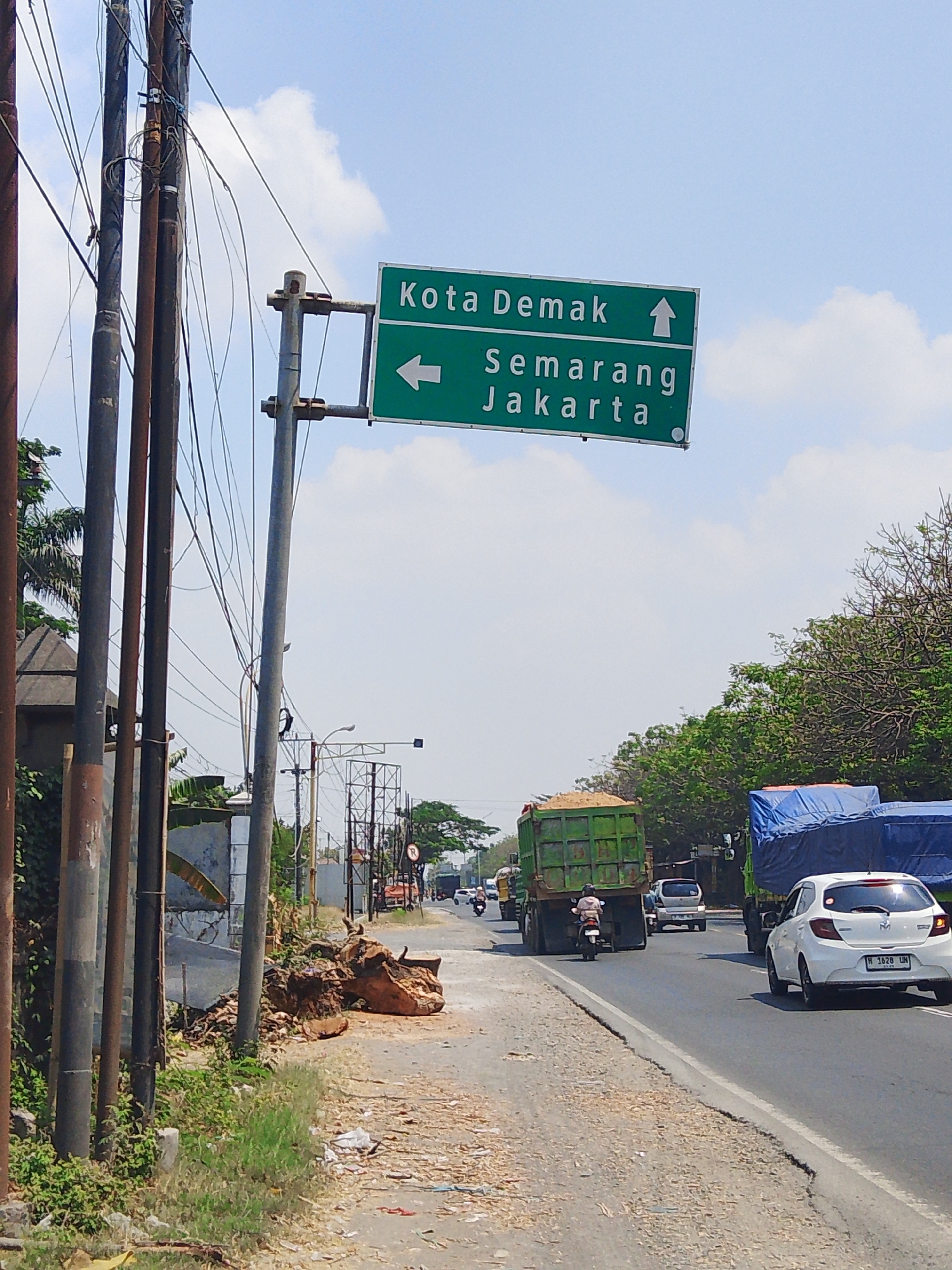 The image depicts a directional signpost on a roadside in Bolo Krajan, Indonesia. The sign, predominantly green with white lettering, indicates directions to "Kota Demak" with an upward arrow, and "Semarang Jakarta" with a leftward arrow. The signpost is supported by a metal pole and is located next to several utility poles with numerous wires.
In the foreground, to the left of the sign, is a pile of debris, including what appears to be a large, weathered tree trunk. The ground along the roadside is covered with gravel and some scattered trash.
The road is a paved, multi-lane thoroughfare. Several vehicles are visible: a white car with license plate "H 1628 UN" is driving on the right lane, a large green truck filled with sand is in the left lane, and other trucks and motorcycles are further down the road. Trees line the right side of the road, and the sky is clear with scattered clouds. The overall scene suggests a typical roadside environment in a developing area, possibly on the outskirts of a city.