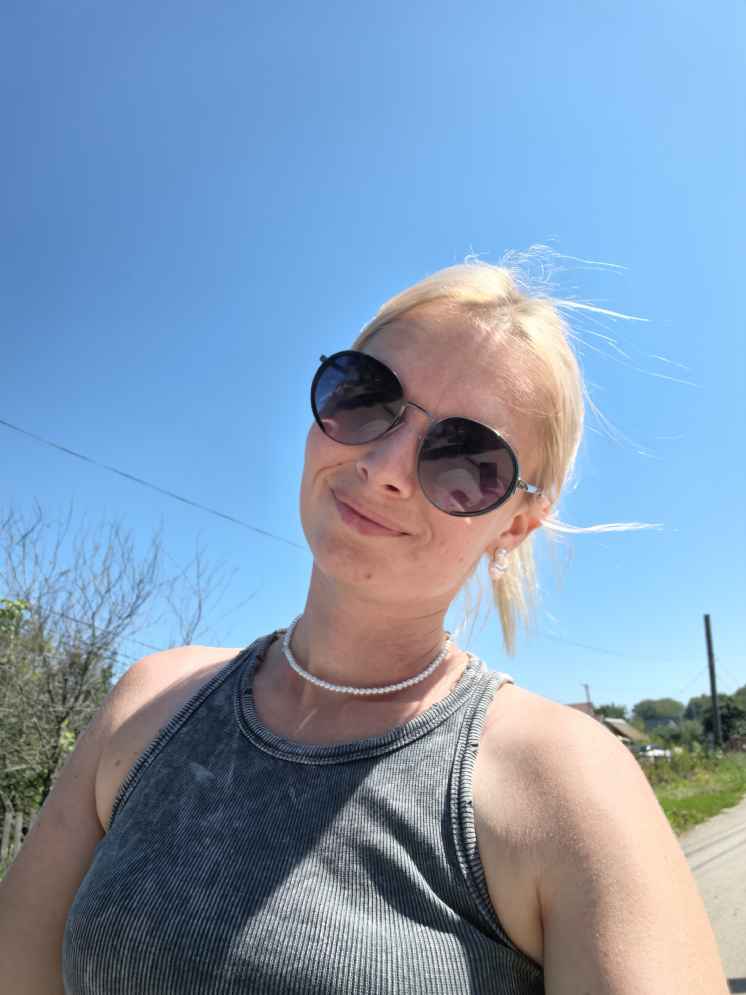 A woman with blonde hair and sunglasses smiles at the camera, holding the camera out to take a selfie. She is wearing a grey tank top and a pearl necklace. The background is a bright blue sky with some trees and a rural setting visible in the distance. The image does not contain any information that would allow for its location to be identified as Sterlitamak, Russia.