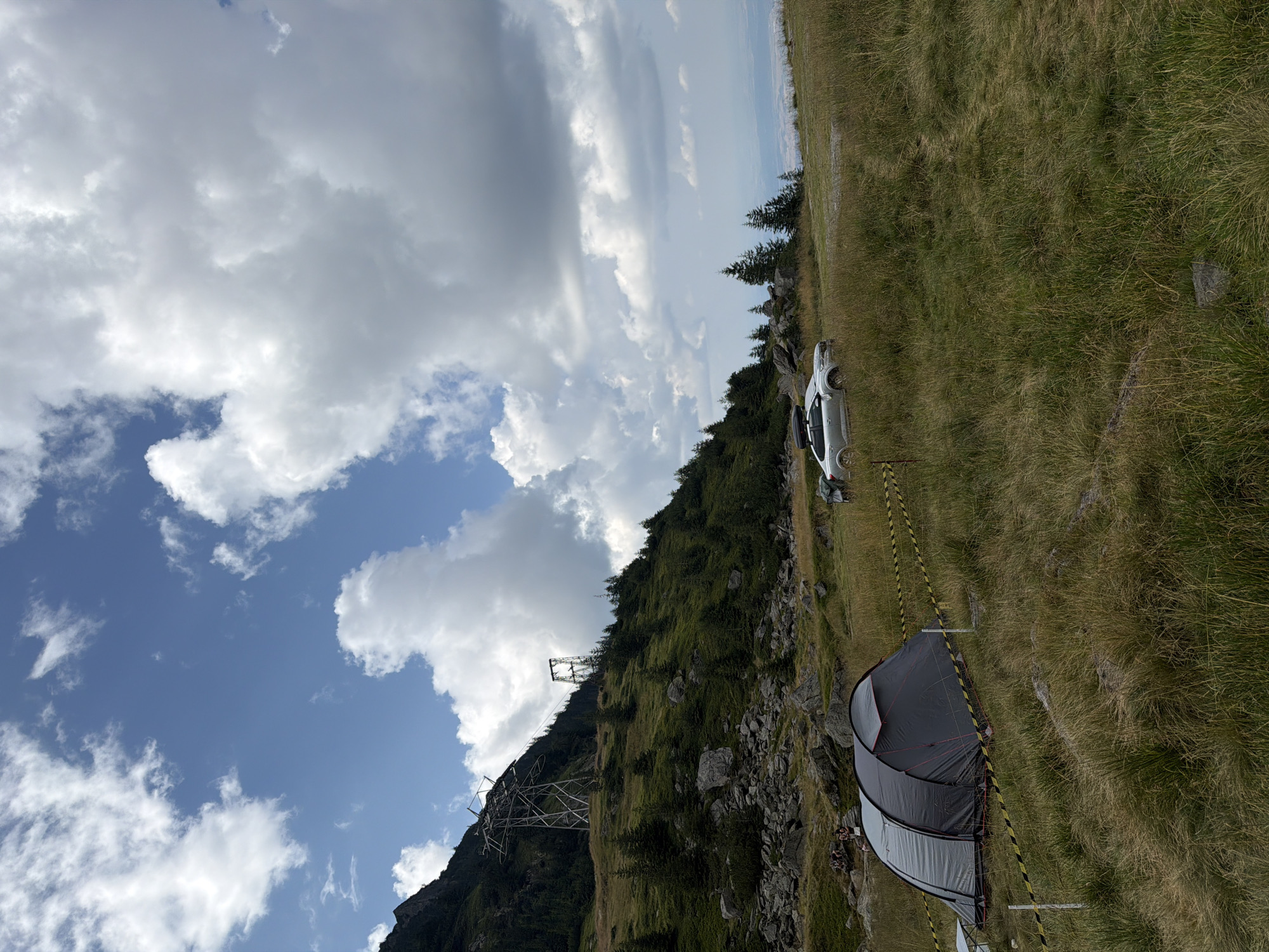 In Cârțișoara, Romania, a mountainous terrain is depicted with a clear sky and scattered clouds. In the foreground, a grey and black tent is set up in a grassy field, cordoned off by yellow and black striped tape. To the right of the tent, a silver SUV with a roof rack is parked. The midground shows a grassy slope dotted with trees and large rocks, leading up to a mountain. A large metal electricity pylon stands prominently on the mountainside. In the background, a distant city or town can be seen. Several people are gathered near the base of the mountain on the left side of the image. The overall scene suggests a camping or recreational activity in a natural, elevated environment.