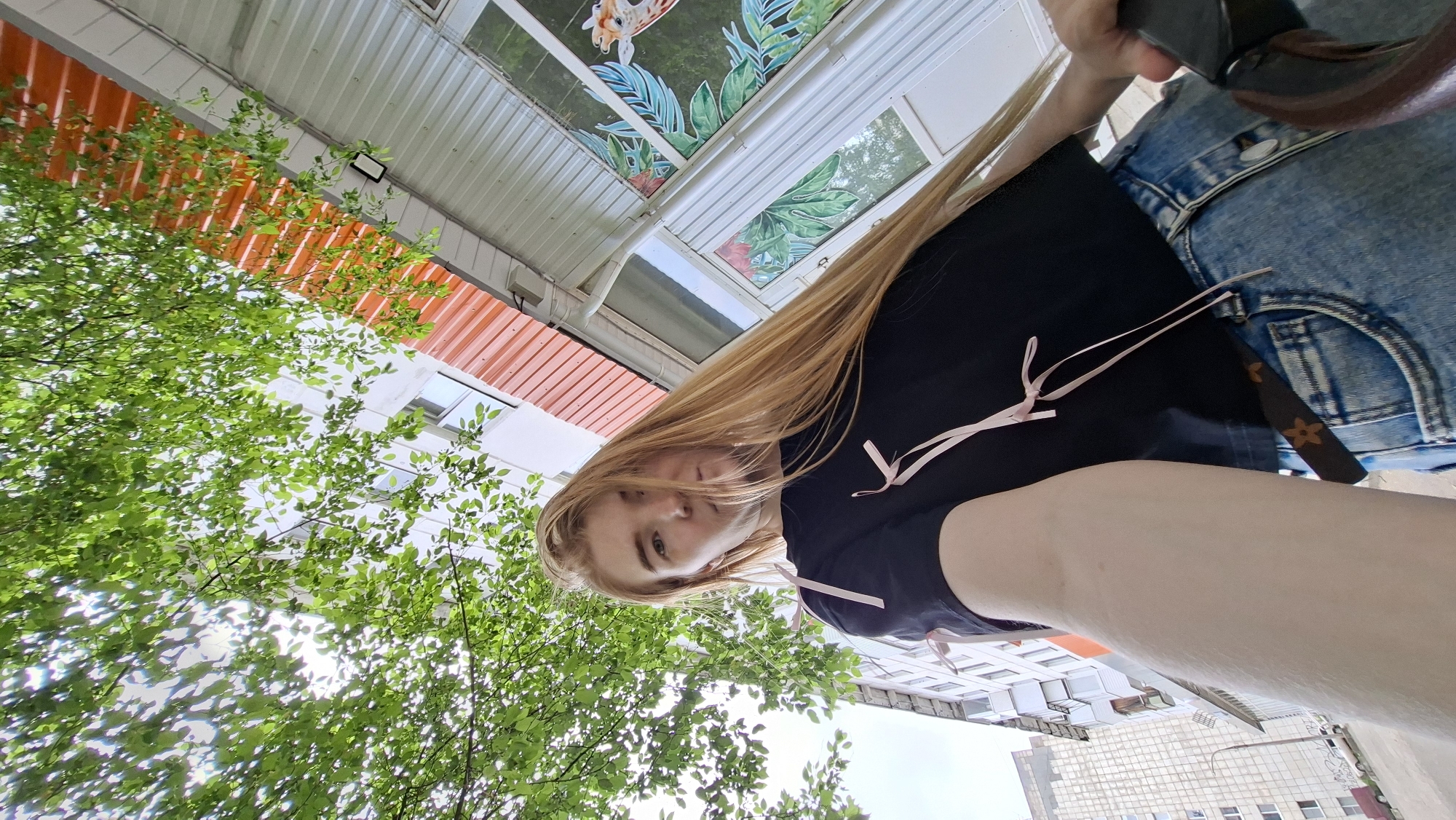A blonde-haired person wearing a black top and blue denim shorts is holding a mobile phone in the upper right of the frame. The person is looking down at the camera. Overhead, there is a roof structure with orange corrugated metal panels and some glass panels decorated with a giraffe design. A tree with green leaves is visible to the left. In the background, a multi-story building can be seen. It can be assumed this image was taken in the city of Chaikovsky, Russia.