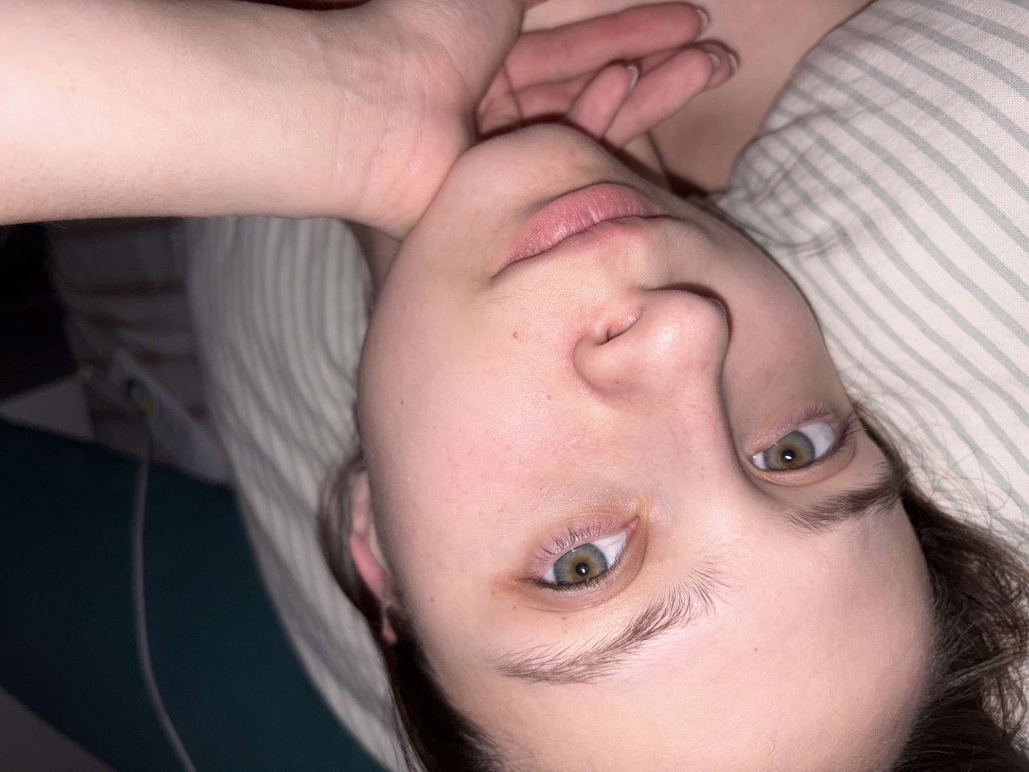 The image shows a person lying down, possibly in a bed, with a striped pillow behind their head. The person is looking upwards and slightly to their right. Their right hand is near their face. Their eyes are hazel. The scene appears to be indoors, likely in a residential setting. No information is available in the image that allows to identify the location in the city of Прикамский, Russia.