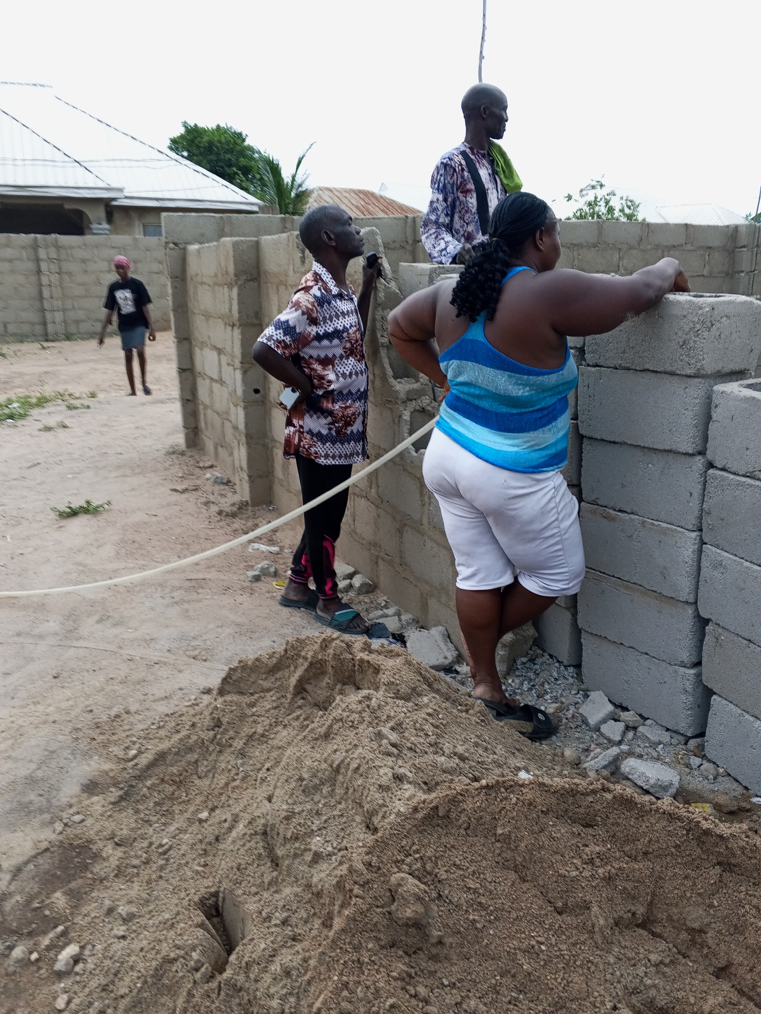 The image shows a construction site in Zango-ndaji, Nigeria. In the foreground is a large pile of sand. A woman wearing a blue and white striped tank top and white shorts stands near a stack of cinder blocks, resting her arm on the blocks. A man in a patterned shirt and black pants is standing behind her, holding a cell phone in his hand and looking upwards. Behind them, another man stands on top of a partially constructed wall wearing a patterned long sleeve shirt, and a green cloth. In the background, a person in a black shirt and dark shorts walks along a dirt path near a low wall. A building with a white roof is visible behind the wall. A white plastic tube runs across the ground in front of the pile of sand.