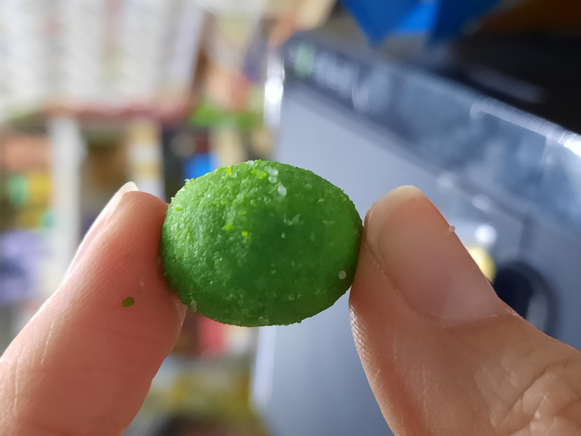 A hand holds a small, round, green confection. The confection is speckled with white crystals. The background is blurred, showing hints of other objects. It is not possible to determine the location as Krasnogorskoye, Russia, from this image.