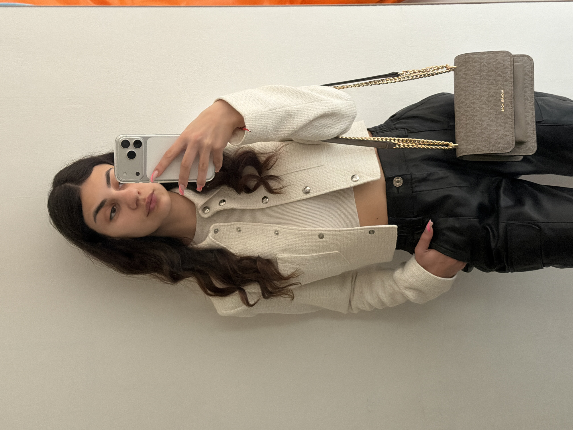 A young woman takes a selfie in what appears to be an indoor environment, possibly a home or apartment. She holds a smartphone in her left hand, obscuring part of her face. The woman has dark hair and is wearing a white tweed jacket over a white top, paired with black leather pants. A Michael Kors purse hangs from a gold chain on her right side. The background is a plain, light-colored wall. No information suggests this image was taken in Gabrovo, Bulgaria.