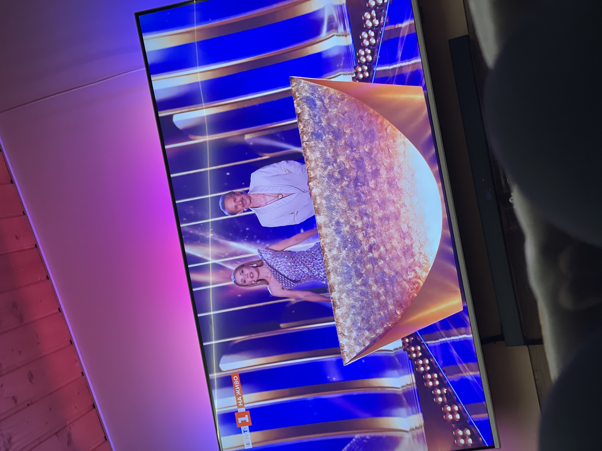 A flat-screen television displays a scene from a broadcast, possibly a competition or awards show. A woman in a sparkly dress and a man in a white suit stand in front of a backdrop of blue and gold horizontal lines and strings of lights. A large, shiny, gold geometric shape partially obscures the scene, suggesting a trophy or award. The "1" logo and the Cyrillic words "НА ЖИВО" (meaning "LIVE") are displayed on the lower left of the screen. The television is located in an indoor setting with a wall painted in shades of pink and purple. The city of Sofia, Bulgaria is likely the location of the TV broadcast.