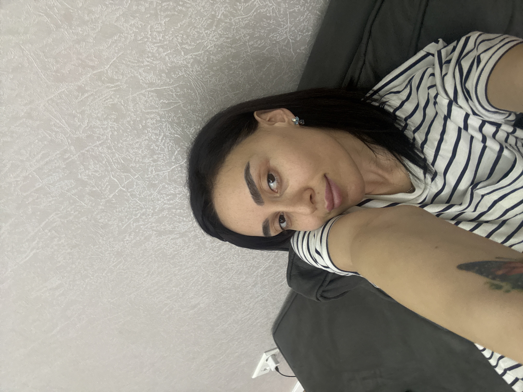 A woman with dark hair, dark eyebrows, and fair skin is lying on a gray couch. She is wearing a white and blue striped shirt. A tattoo is visible on her right arm. The background is a light gray wall with a textured pattern. A white electrical outlet with a plug is visible near the bottom of the image. The location cannot be determined from the image.