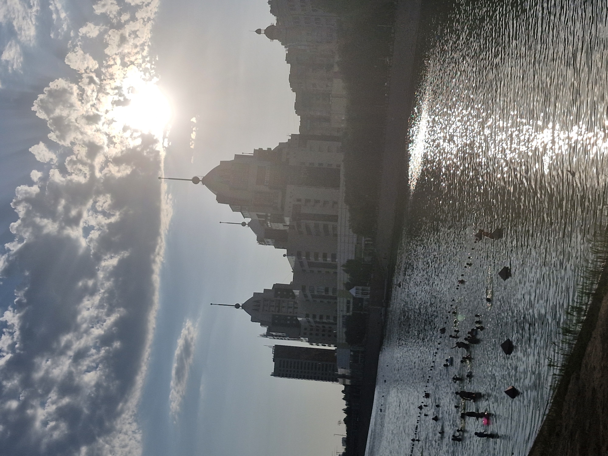 The image captures a sunlit day in Astana, Kazakhstan, with the Ishim River flowing through the city. The sun is high in the sky, partially obscured by dramatic clouds, casting a bright glare on the water's surface. Silhouetted against the bright sky are several modern, distinct buildings, some featuring unique architectural elements like spires and stepped facades. The river is populated by numerous people swimming and wading, creating a lively scene. Several buoys or markers are visible in the water, possibly delineating swimming areas. The foreground shows a grassy embankment. The overall impression is one of a vibrant urban recreational area on a clear day.