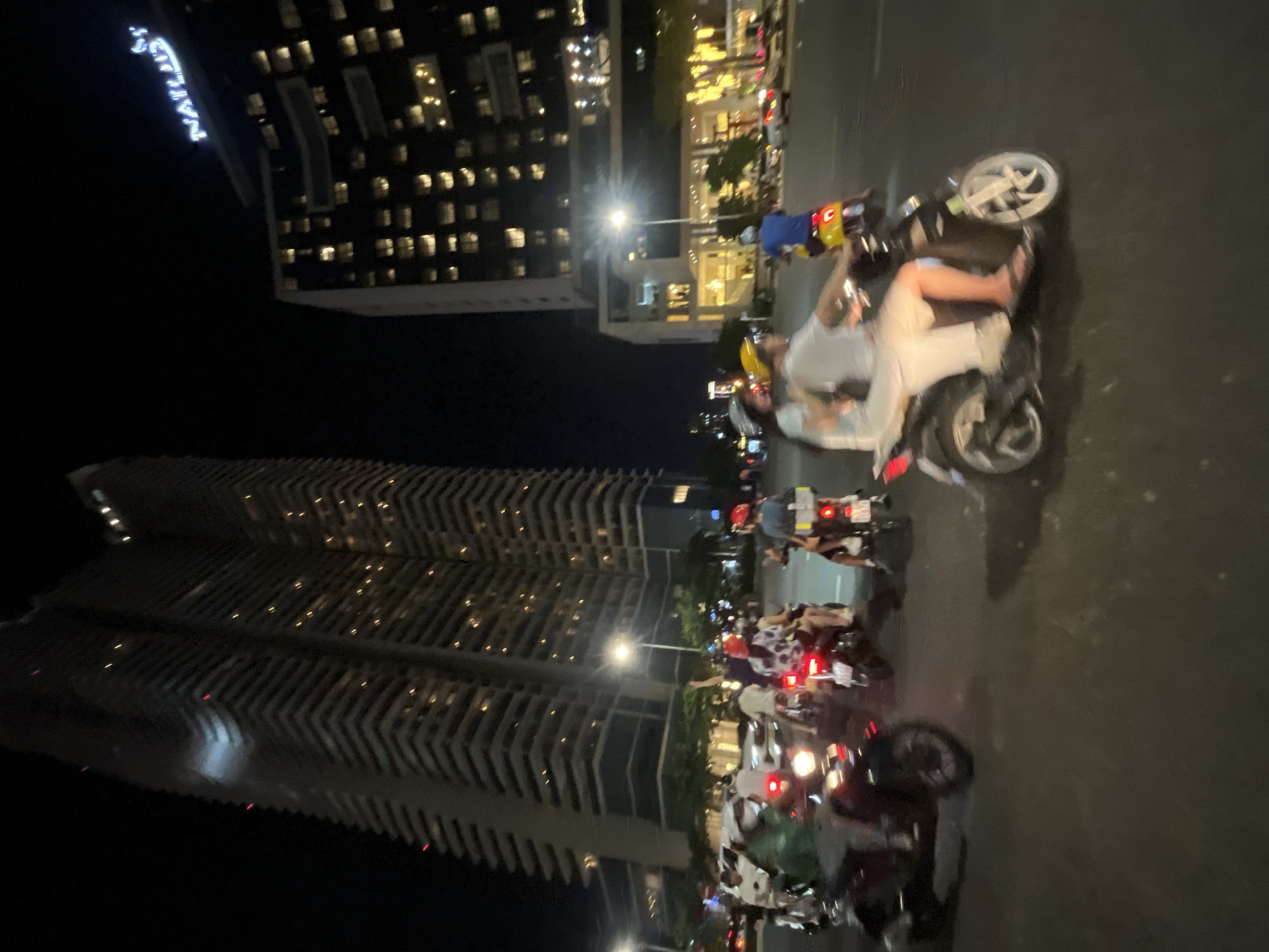 The image depicts a nighttime street scene in Đà Nẵng, Vietnam, featuring a procession of motorbikes. Two prominent skyscrapers dominate the background. The left skyscraper is tall and modern, with a facade of many windows and balconies. The right skyscraper, partially obscured, displays a lit sign that reads "NATURIST". The street is illuminated by streetlights and the lights from the buildings and motorbikes.
In the foreground and middle ground, several motorbikes are in motion. The riders are wearing helmets and casual clothing. Some motorbikes have passengers. The motorbikes are of various types, with some appearing to be scooters and others more like traditional motorcycles. The riders' faces are not clearly distinguishable due to the motion blur and low light conditions. The overall atmosphere suggests a bustling urban environment at night. The road surface appears wet, possibly from recent rain, reflecting the lights.