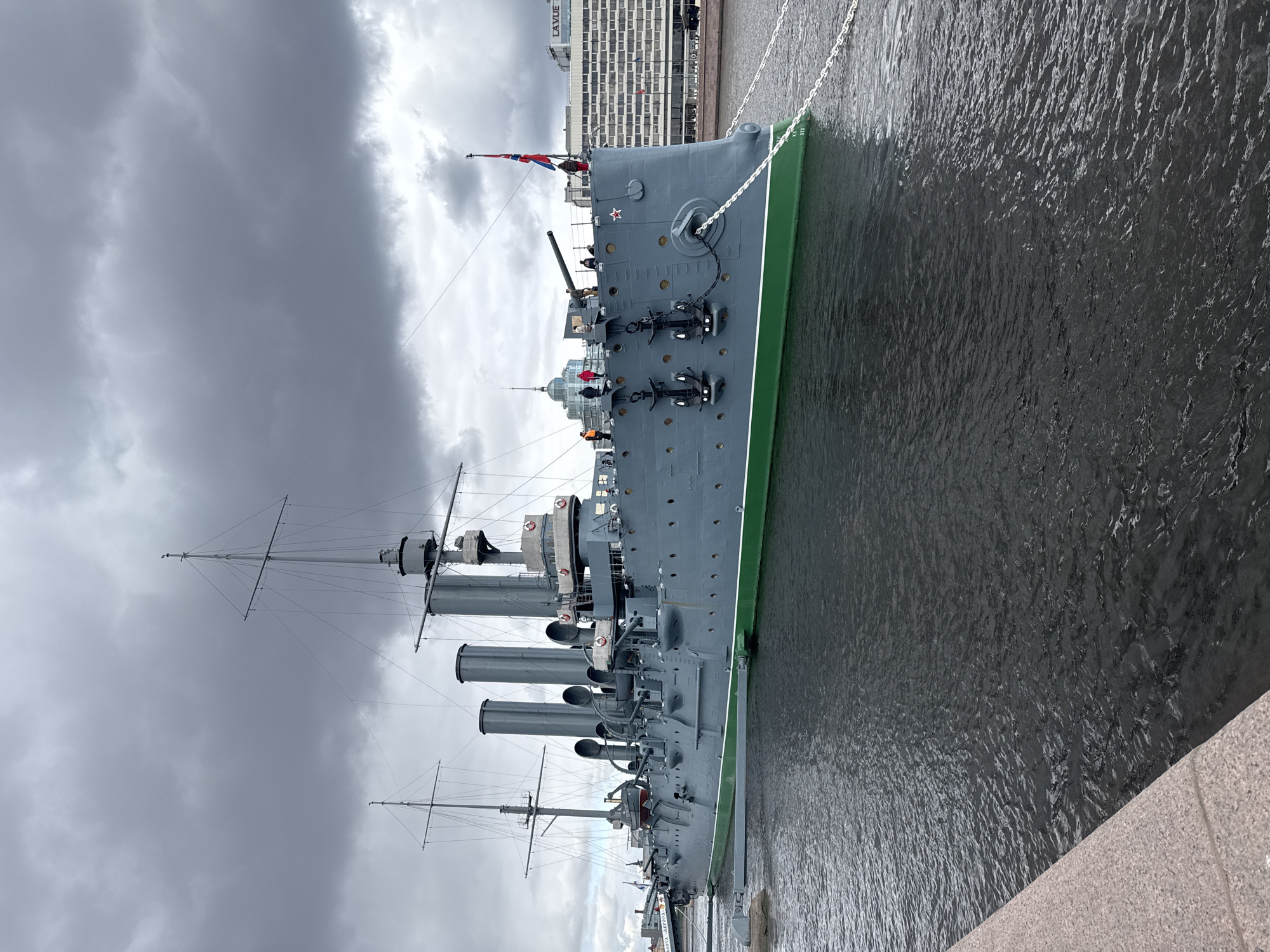 The image shows the Cruiser Aurora, a retired Russian warship and museum ship, docked in the Neva River in Saint Petersburg, Russia. The ship is painted in gray with a green waterline stripe. It has multiple smokestacks, masts, and gun turrets. A Soviet flag is visible at the stern. The ship is secured by mooring lines to the embankment. The Neva River is murky and reflects the cloudy sky above. In the background, modern buildings are visible along the riverbank. The sky is overcast with dark clouds. A stone embankment runs along the foreground of the image.
