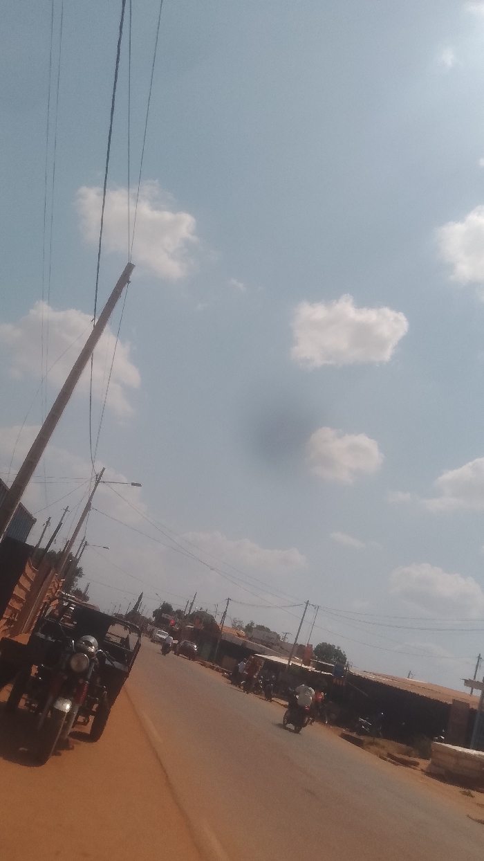 The image depicts a street scene in Korhogo, Côte d'Ivoire, under a clear blue sky with scattered white cumulus clouds. A pronounced dark circular artifact is visible in the upper sky, likely a lens smudge.
In the immediate foreground, on the left, a reddish-brown dirt shoulder runs alongside an asphalt road. Two motorcycles are parked on this shoulder, one with a side-mounted cargo bed (a motortricycle), facing generally away from the viewer. Utility poles with multiple overhead wires ascend diagonally from the left side of the frame towards the sky. Some poles also feature street lighting fixtures.
The asphalt road, angled from the lower center towards the upper right, serves as the main thoroughfare. Traffic is visible, primarily consisting of motorcycles and at least one car in the distance. Several individuals are seen riding motorcycles. One motorcyclist in the mid-ground, wearing a white shirt, is traveling away from the viewer on the right side of the road. Other individuals are present further down the road, some possibly on motorcycles, others potentially pedestrians near the roadside structures.
Along the right side of the road, a series of structures with dark, possibly corrugated metal roofs are present, indicative of shops or market stalls. Beyond these structures and the road, trees and other distant buildings are visible under the horizon line. The scene suggests a bustling urban or semi-urban environment.