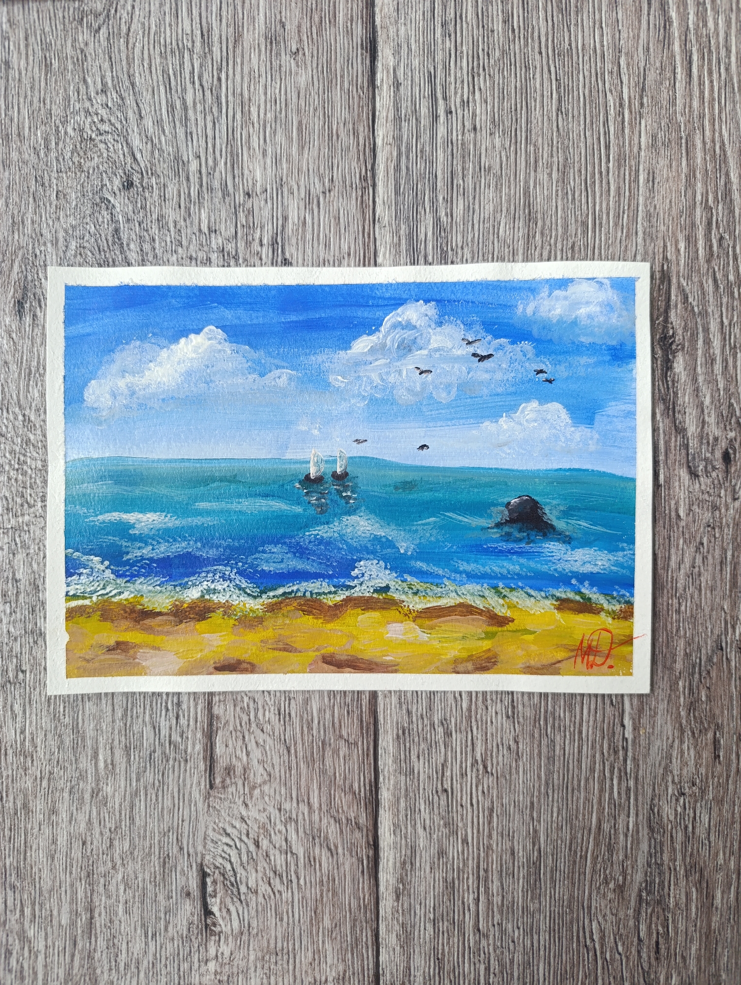 A horizontal, rectangular acrylic painting on paper depicts a seascape with a beach, ocean, and sky. The background is a rustic wooden surface. The sky is a vibrant blue with scattered, fluffy white clouds. Several black birds are in flight across the sky. The ocean is a deep turquoise with white-capped waves crashing onto the shore. Two sailboats are visible in the distance. In the mid-ground of the ocean, a dark, rocky outcropping emerges from the water. The foreground is a sandy beach with patches of brown and yellow. A signature in red paint, "MD", is visible in the bottom right corner of the painting.