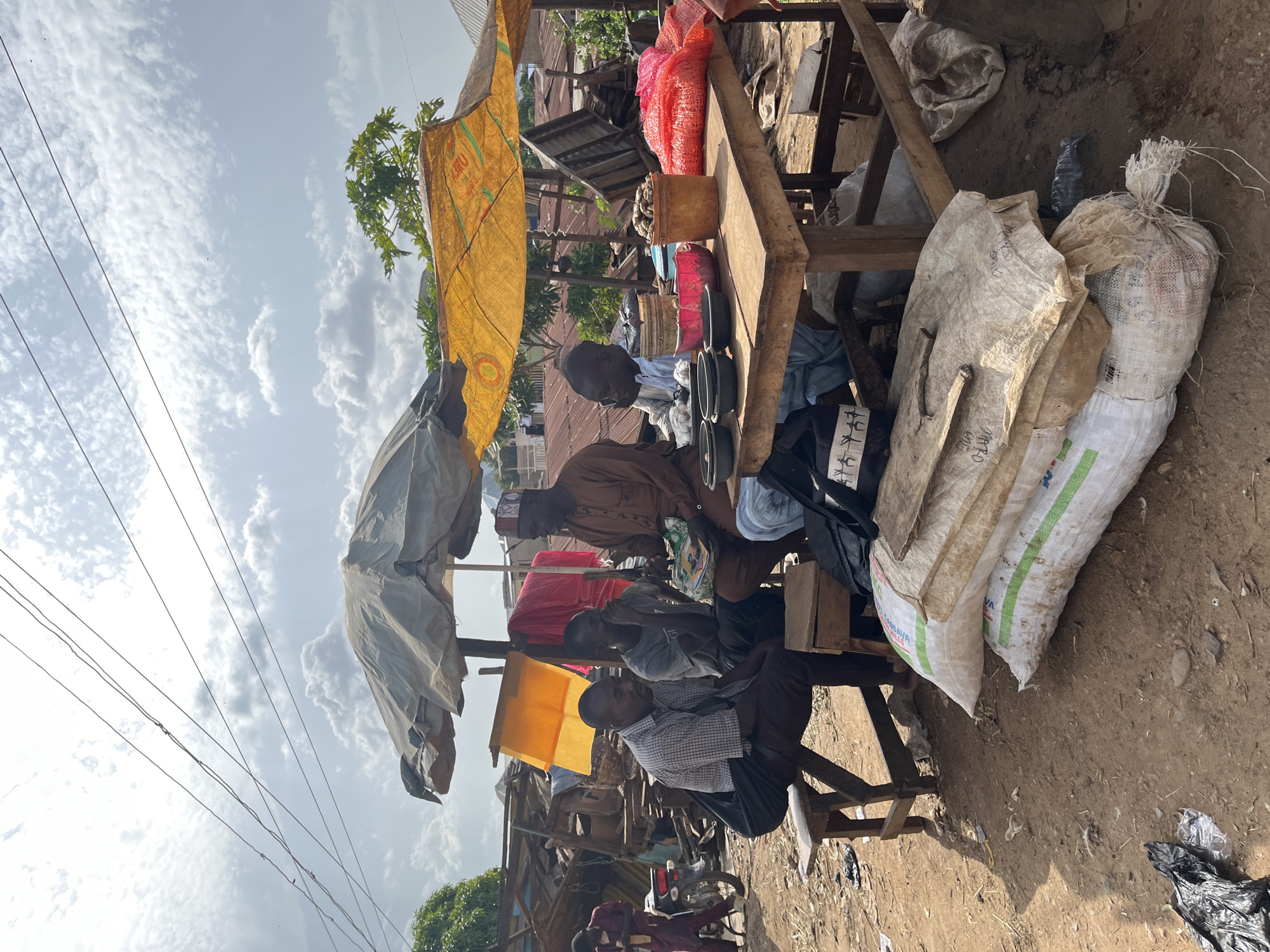 The image depicts an outdoor market scene in Jos, Nigeria, under a partly cloudy sky. Several men are gathered around a wooden table laden with goods. Two men are seated on a bench in the foreground, with one looking towards the table. Another man in a brown tunic and cap is seated at the table, holding a book or papers. A fourth man, wearing a light-colored shirt, is also at the table, appearing to be engaged in conversation.
On the table, there are several dark bowls, a pink bowl, a bucket filled with nuts or roots, and a pile of red netting containing produce, possibly onions or potatoes. Several large sacks are stacked on the ground in front of the table. In the background, makeshift stalls constructed from corrugated metal and wood are visible, along with trees and more distant buildings. A motorcycle and a person in maroon clothing are seen to the left, suggesting ongoing activity in the market. Power lines crisscross the sky above the scene. The overall impression is of a bustling, informal marketplace.