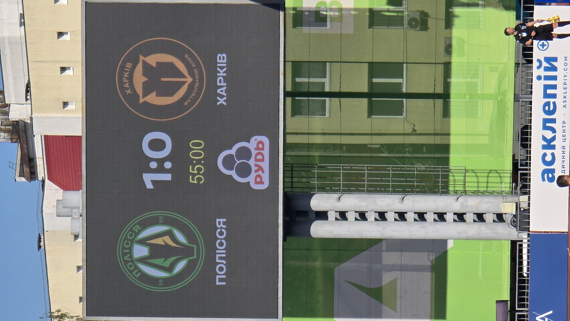 The image captures a scoreboard at a football stadium, displaying a match between "POLISSYA" and "KHARKIV". The score is 1:0, and the game is at the 55:00 mark. The logos of both teams are visible. Below the team names, a sponsor's logo, "РУДБ", is displayed. The scoreboard is set against a backdrop of stadium architecture and a bright blue sky. In the foreground, partially visible, are stadium structures and a green backdrop. At the bottom of the image, a man in a referee's uniform, holding a yellow card, is standing, and another man is partially visible in the lower left corner. The image appears to be taken from a distance, focusing on the scoreboard. This scene is likely at the Tsentralnyi Stadion in Zhytomyr, Ukraine, which is home to the FC Polissya Zhytomyr football club.