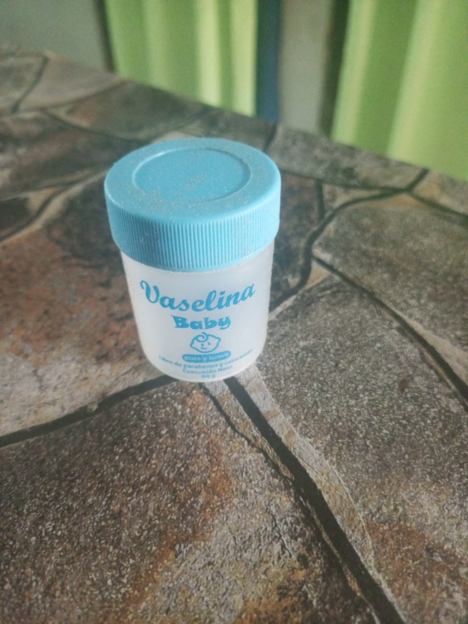 A small, cylindrical jar of Vaselina Baby lotion sits on a rough, stone-like surface. The jar is white with a light blue lid. The word "Vaselina" is printed in a stylized font above "Baby" with a small baby face icon. Below this, the words "pura y suave" and "Libre de parabenos y colorantes. Contenido Neto 50g" are printed in smaller text. The background is blurred, showing indistinct green and white vertical stripes. The scene is an indoor setting. Specific location within Juan Griego, Venezuela cannot be determined from this image.