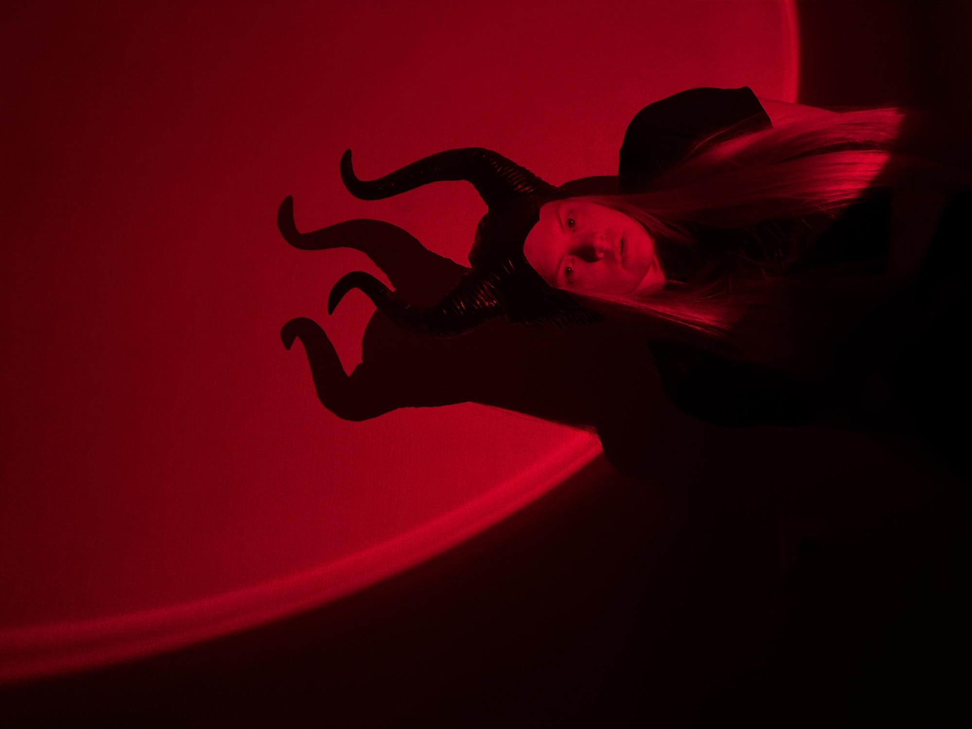 The image captures a dramatic, dark scene illuminated predominantly by a striking red light. The central subject is a person, likely female, positioned horizontally across the frame, looking directly towards the viewer. She has long, straight blonde hair that cascades to her left, catching the red light and appearing vibrant against the dark background.
She is wearing a dark costume that includes prominent, textured black horns protruding from her head, resembling those of a demon or a character like Maleficent. These horns cast a large, distinct silhouette on the red surface behind her. Her face is softly illuminated by the red light, revealing a neutral yet intense expression with her eyes open.
The setting appears to be an indoor space or studio, devoid of environmental cues, where a deep red light source bathes the background. There are noticeable patterns of light and shadow on the red surface, including a curved, brighter line of red light stretching across the lower-left portion of the frame, suggesting a projected light effect or a specific type of lamp. The right side of the image, beyond the subject, recedes into deep shadow, creating a stark contrast with the illuminated areas. The overall impression is one of theatricality and mystery. There is no visible text in the image.