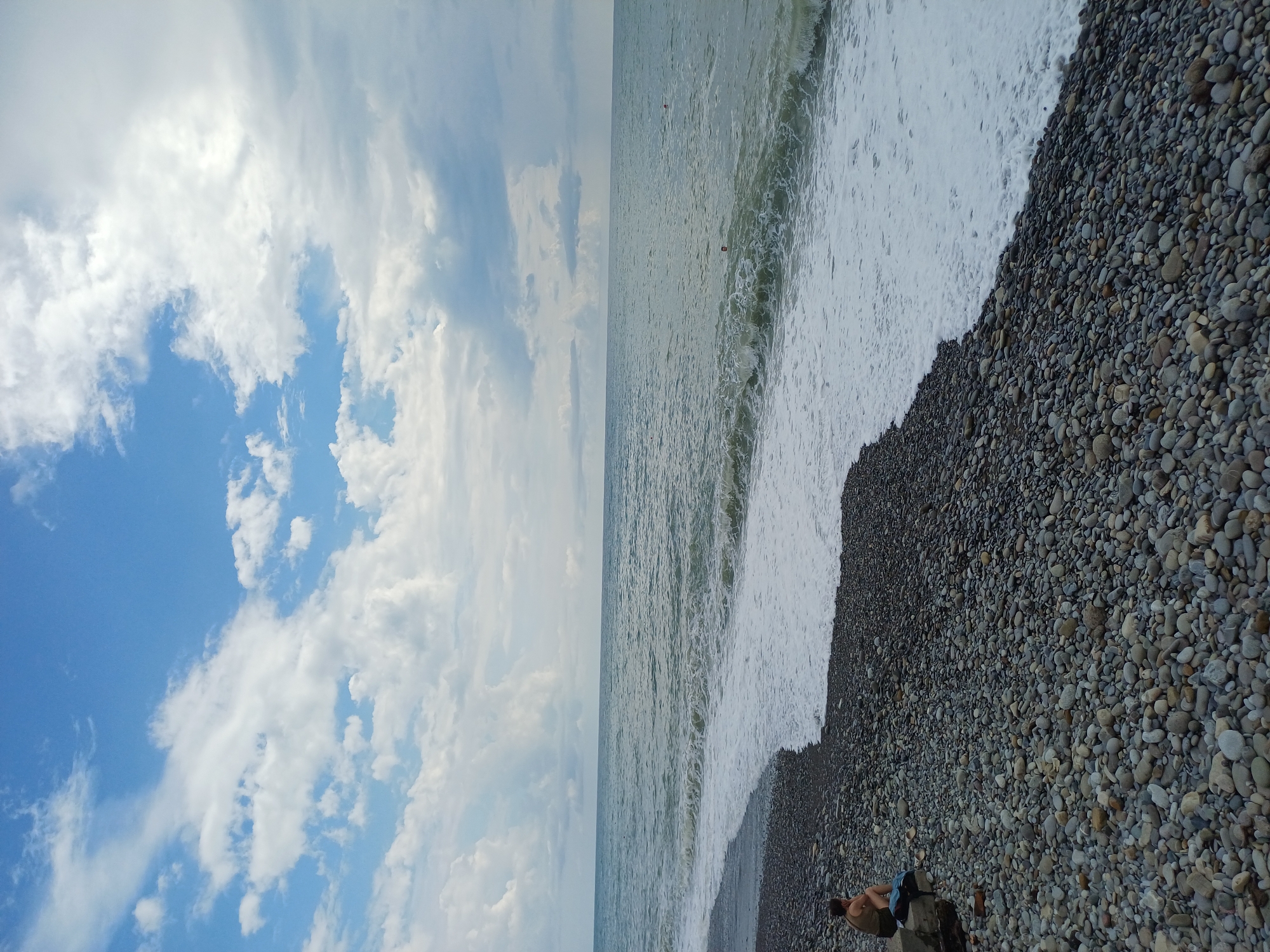 The image depicts a pebble beach scene on the coast of Sochi, Russia. The sky is filled with a mix of white, fluffy clouds and some grayer, more diffused clouds, against a bright blue backdrop. The sea is a muted greenish-blue, with moderate waves creating a white foamy line as they break on the shore.
In the lower left quadrant of the image, a person, appearing to be male and wearing a dark t-shirt and shorts, is seated on a large, dark rock. Next to him, a dark blue bag is placed on the rock. The foreground is dominated by a dense covering of smooth, gray, and multicolored pebbles. The waterline forms a distinct curve across the frame, separating the wet, darker pebbles closest to the sea from the dry, lighter pebbles further up the beach.
In the distance, a few small, red buoys are visible in the sea. The overall impression is a quiet, perhaps slightly overcast day at the beach.