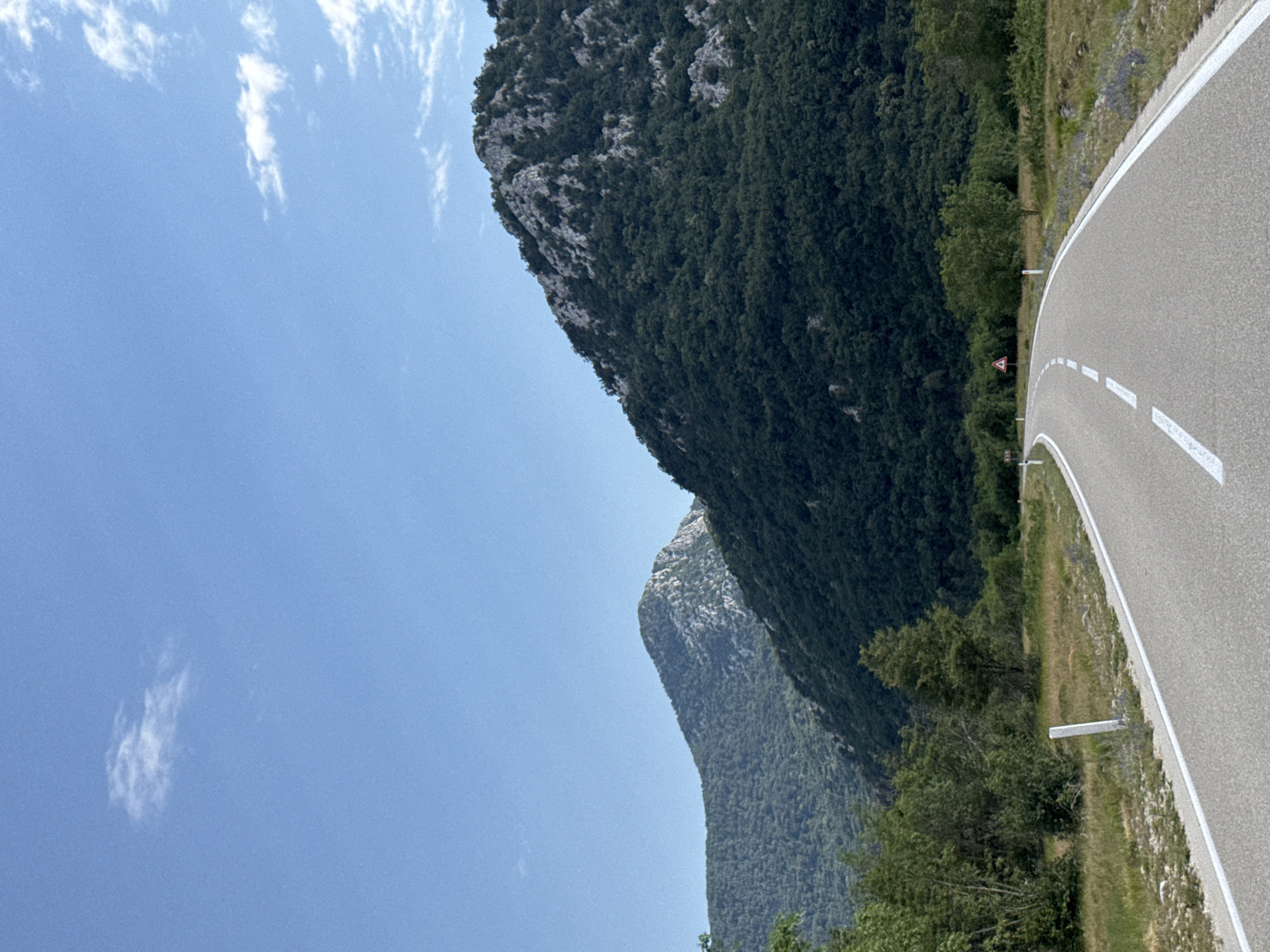 The image captures a scenic view of a winding road leading through a mountainous landscape. The road, paved and marked with white lines, curves gently to the left, disappearing into the distance. On either side of the road, the terrain is covered with green vegetation, including trees and grasses. In the background, majestic mountains rise, their slopes densely forested. The sky above is a clear blue, with a few wispy white clouds scattered across it. The overall impression is one of natural beauty and tranquility. The location is specified as the city of Dragalj, Montenegro. There are no people or animals visible in the image. Essential details of the scene include the road, mountains, vegetation, and sky.