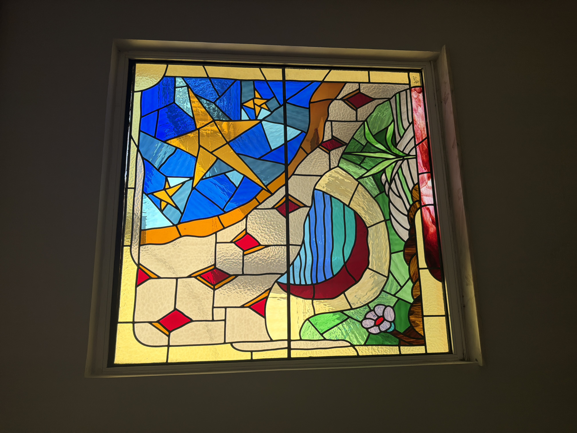 The image shows a stained glass window set into a wall. The window is rectangular and framed by a white border. The stained glass depicts a stylized landscape with celestial elements. In the upper portion, a bright yellow star is prominent against a background of various shades of blue, representing the night sky. There are other smaller stars and geometric shapes in blue and yellow.
The lower part of the window features a crescent moon shape in deep red, filled with wavy lines of blue and green, suggesting water. To the left of the moon, there is a single, stylized flower with white petals and a red center, growing from a brown stem and green leaves. The ground around the flower and the moon is depicted with patches of green and earth tones, interspersed with white and grey abstract shapes. There are also red diamond-shaped pieces of glass scattered throughout the scene. A thin wooden windowsill is visible below the stained glass window. The surrounding wall is painted a dark, neutral color, creating a contrast with the illuminated window. The lighting suggests the image was taken indoors, possibly during the day as light passes through the stained glass. The location in Arcahueja, Spain, is not evident from the visual information provided in the image.