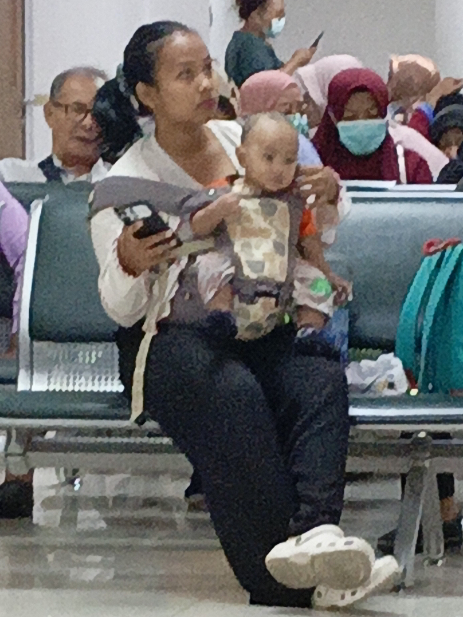In a waiting area in East Jakarta, Indonesia, a woman carries a baby in a carrier while holding a smartphone. She wears a light-colored top and dark pants. The baby is dressed in a camouflage-patterned outfit. Other individuals are visible in the background, some wearing face masks. The environment appears to be indoors, possibly a transportation hub or public facility, with rows of seating. A teal backpack rests on a seat to the right. The lighting is moderate.