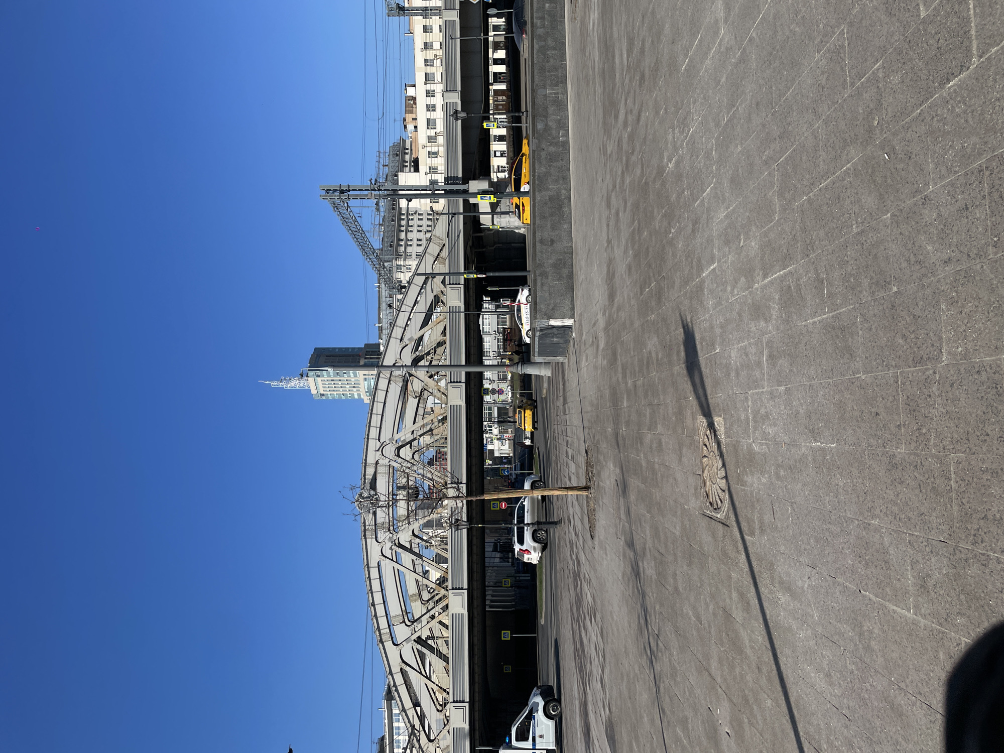 The image shows a cityscape in Moscow, Russia. A steel arch bridge dominates the view, with railway tracks and overhead lines running across it. Under the bridge, there's a road with a white car and a yellow taxi parked along the side. A street lamp stands near a young tree, its bare branches reaching out. In the background, tall buildings rise against a clear blue sky. The foreground is a paved area, its surface textured with interlocking stones. A circular manhole cover sits in the paving, casting a shadow.