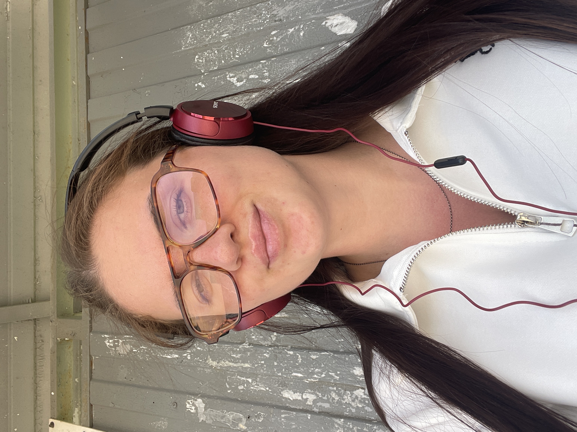 A woman is seen in a selfie, turned to the left in the frame. She wears headphones and glasses, and a white zipper jacket. The background consists of a grey, weathered surface, possibly a wall or floor. No specific details point to Molokovo, Russia.